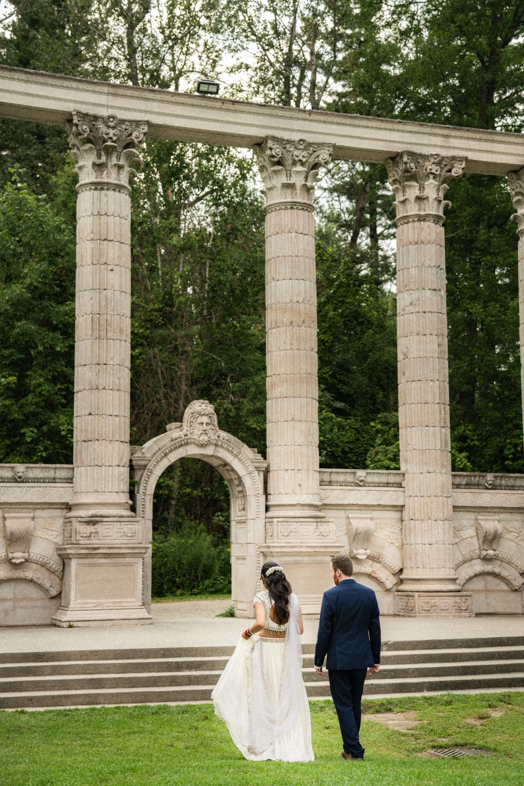 Bride and groom walk hand in hand toward the Greek Theatre columns and carved arch., by AD Photography