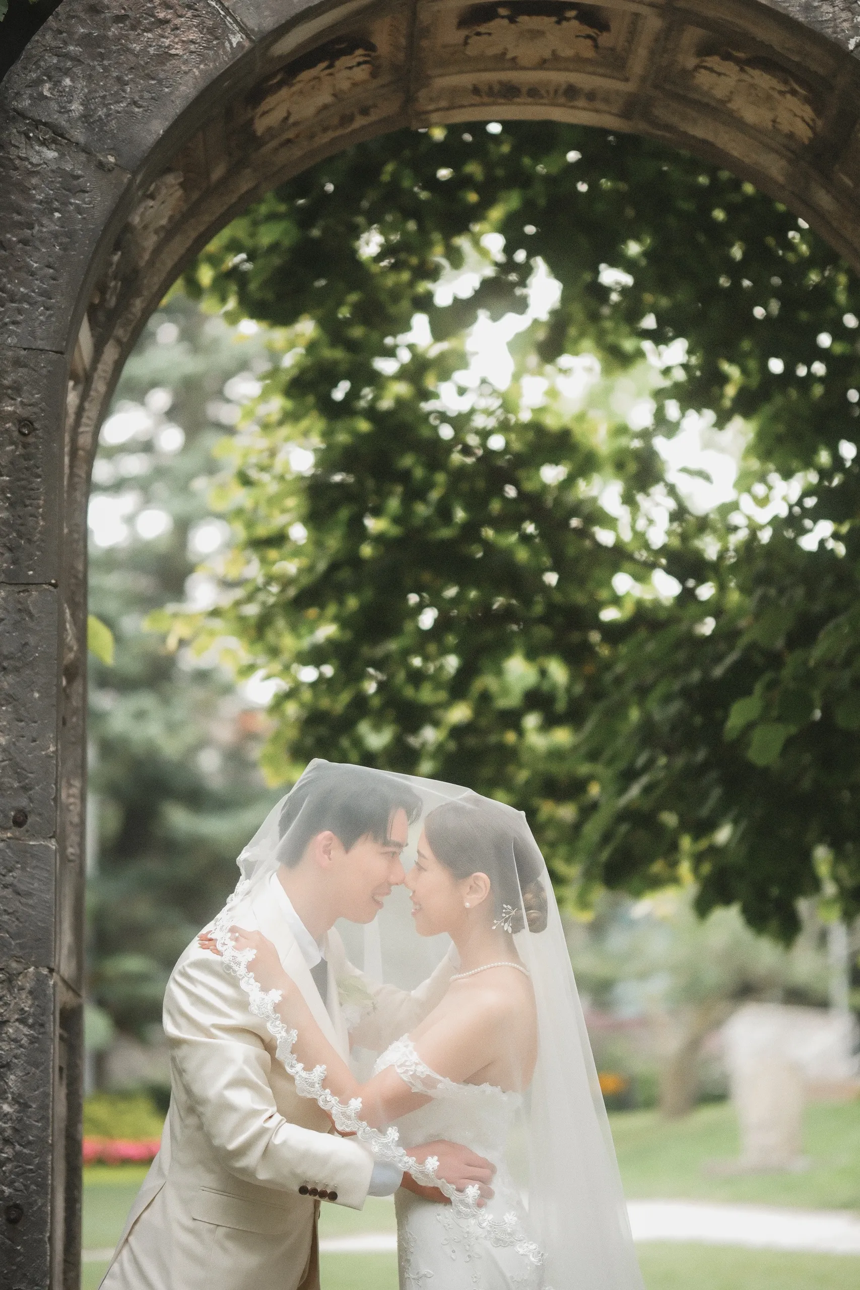 Josie and Nelson sharing her veil beneath a carved stone arch at Guild Park, Scarborough, by AD Photography