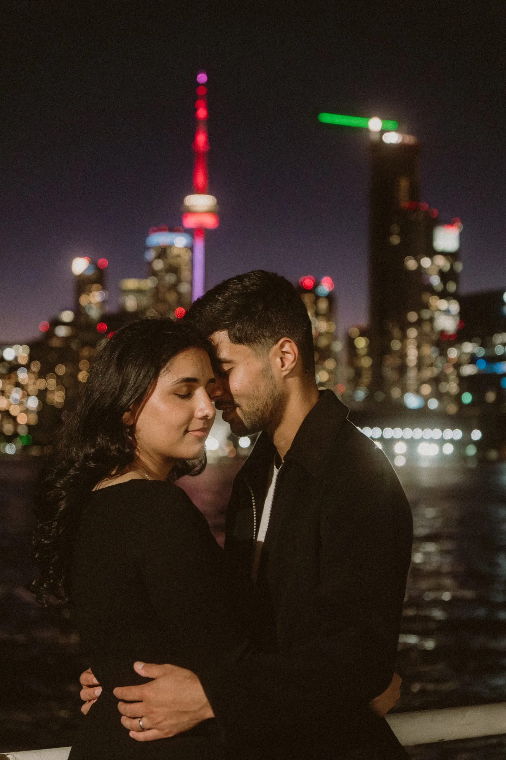 Foreheads together at night with the CN Tower glowing between the couple at Polson Pier, by AD Photography