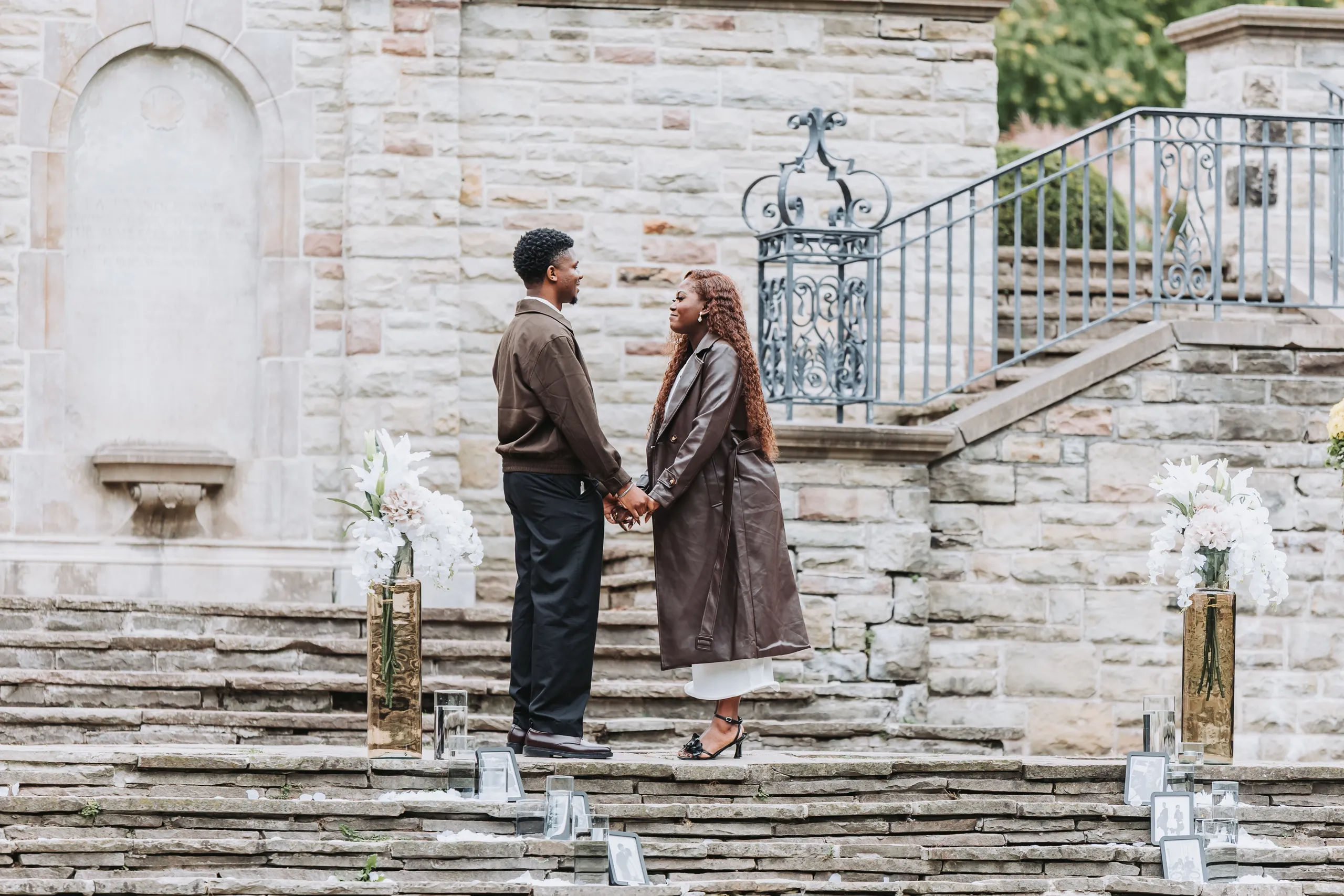 Richie and Aimee holding hands on the terrace steps, white lilies in gold vases on either side, by AD Photography