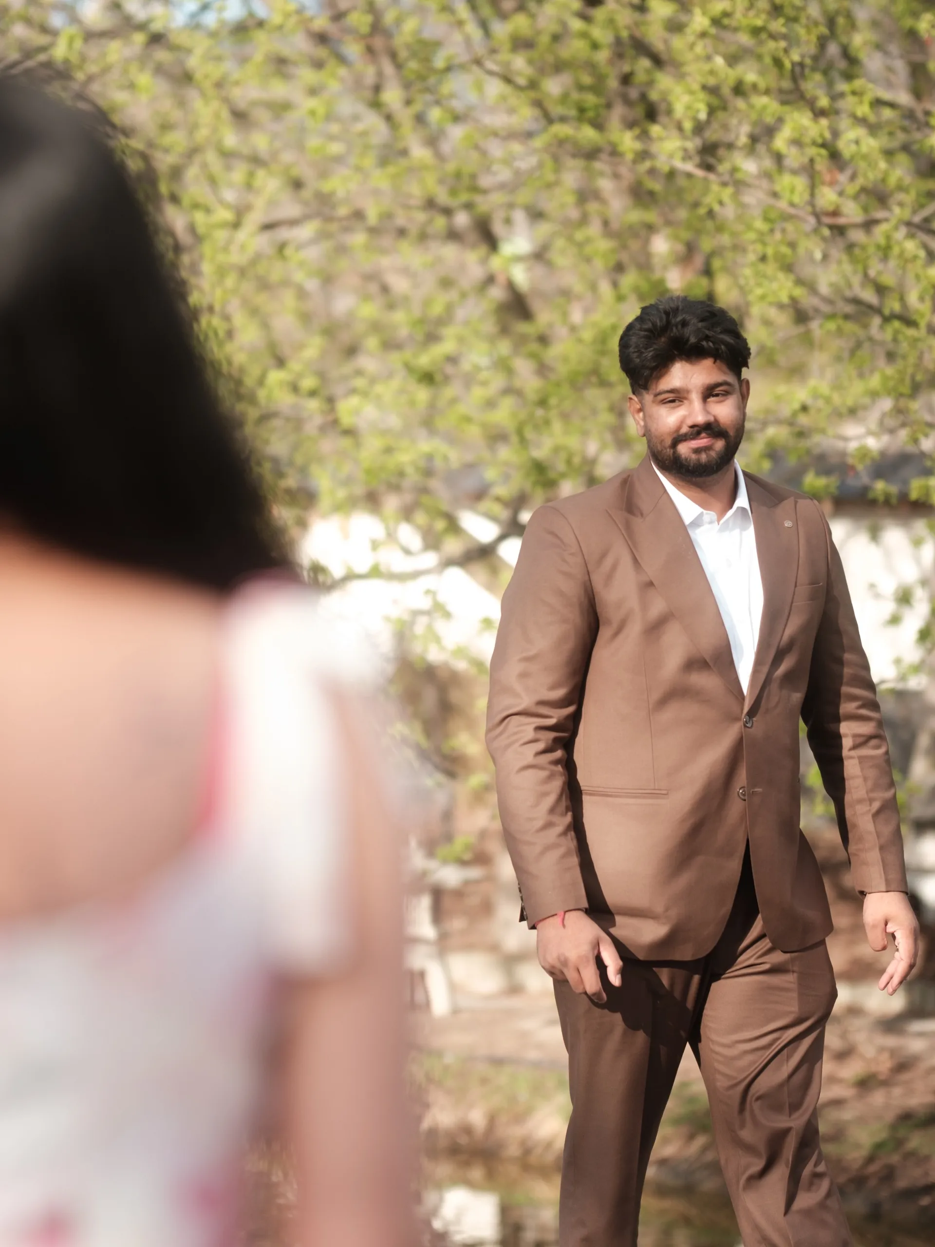 Kamal crossing the garden path toward Mehak in his tan suit, spring branches overhead, by AD Photography