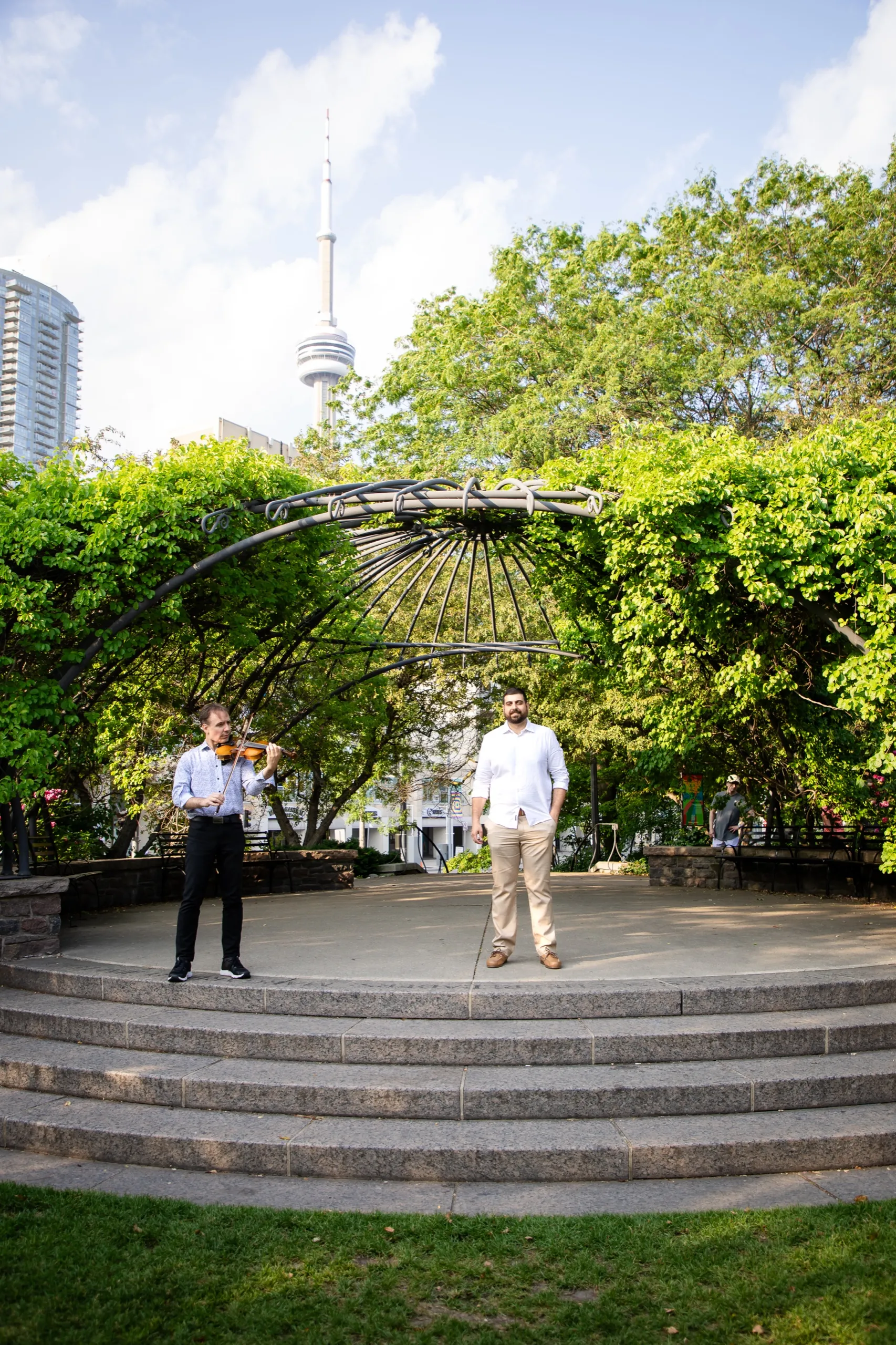 A violinist and the groom to be waiting under the iron pergola at the Toronto Music Garden before the proposal, by AD Photography