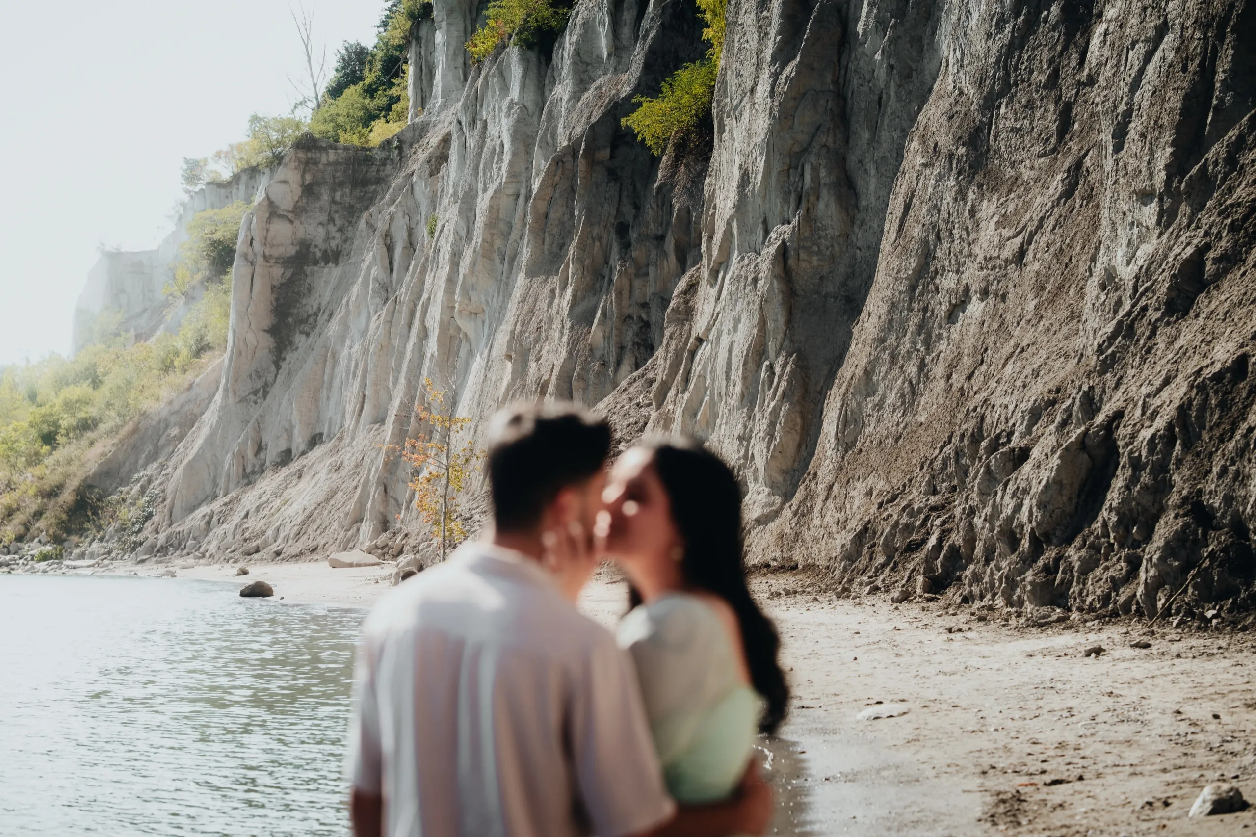 Soft focus kiss at the shoreline with the Scarborough Bluffs razor sharp behind the couple, by AD Photography