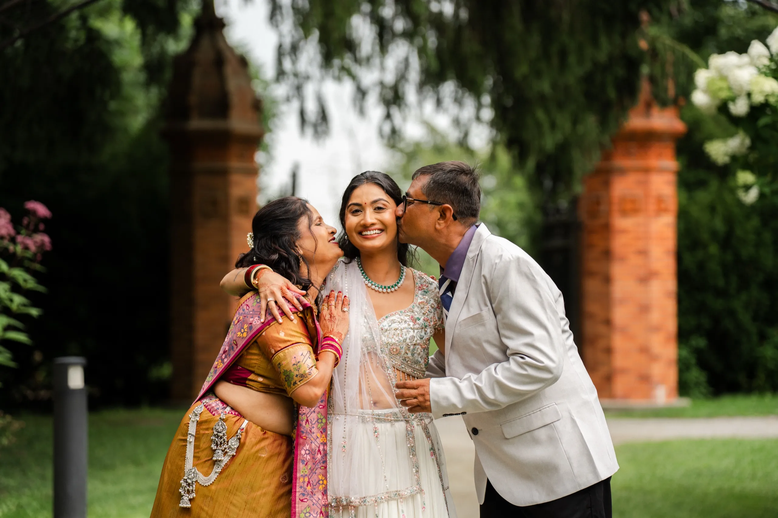 Nidhi's parents kiss her cheeks at once, her bindi and emerald necklace catching the light., by AD Photography