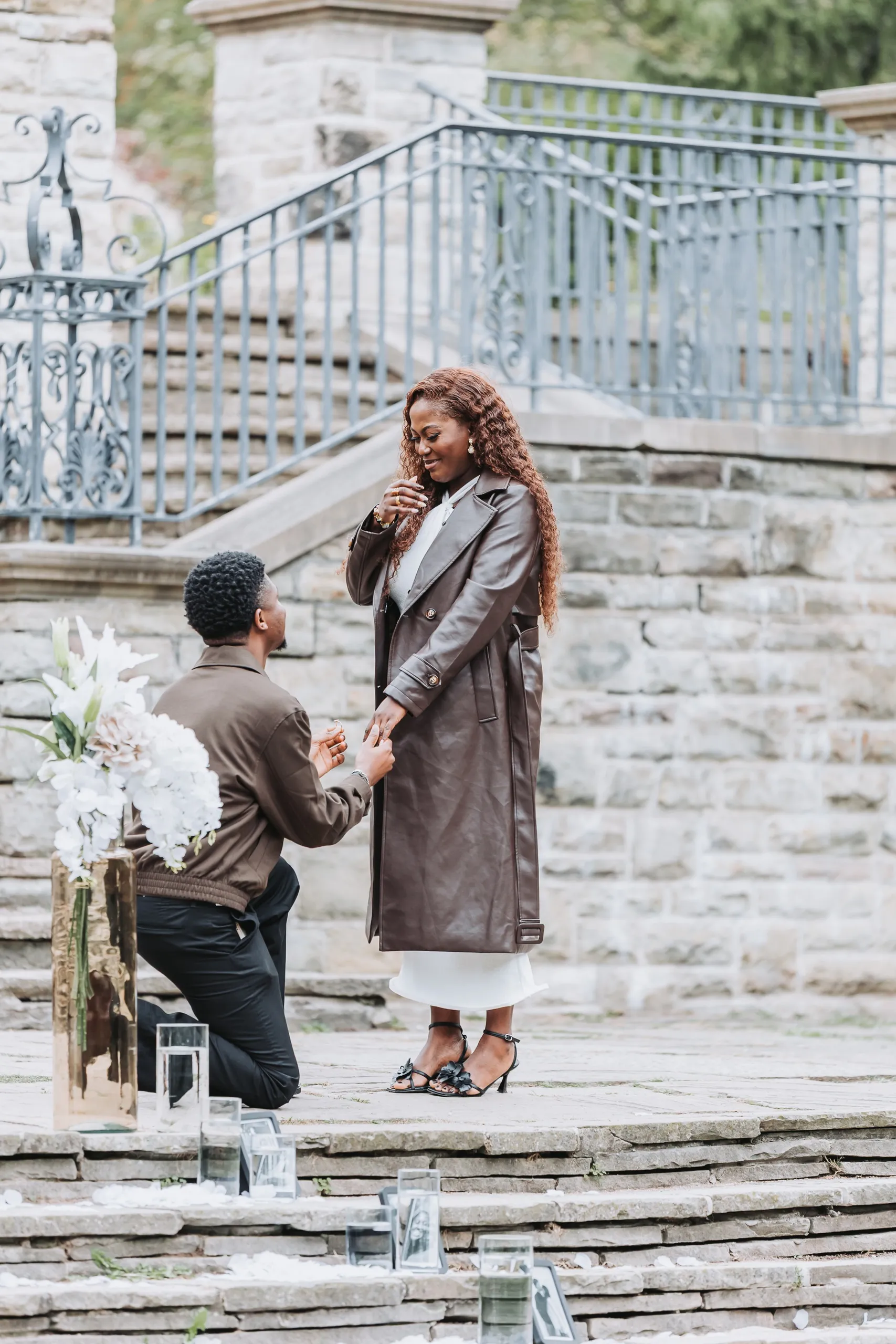 Richie on one knee holding Aimee's hand as she laughs in shock beside the white lilies, by AD Photography