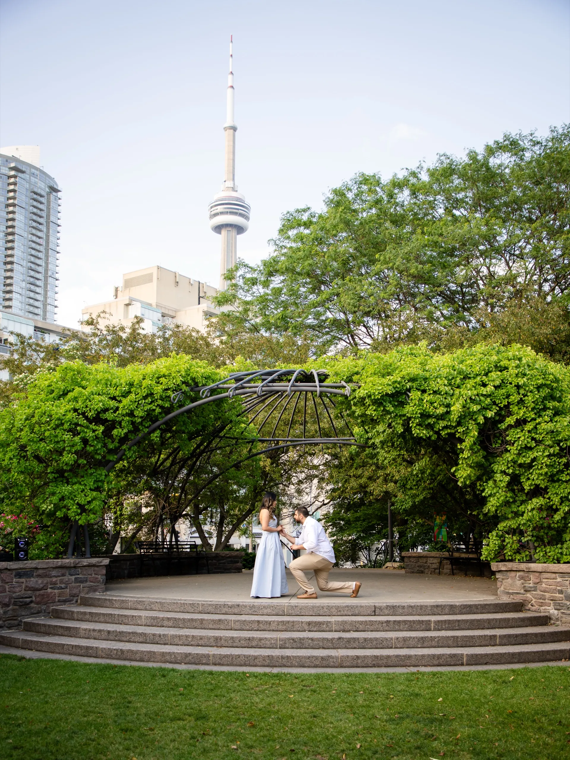 Mike on one knee proposing to Margarita under the pergola at the Toronto Music Garden, with the CN Tower rising behind them, by AD Photography