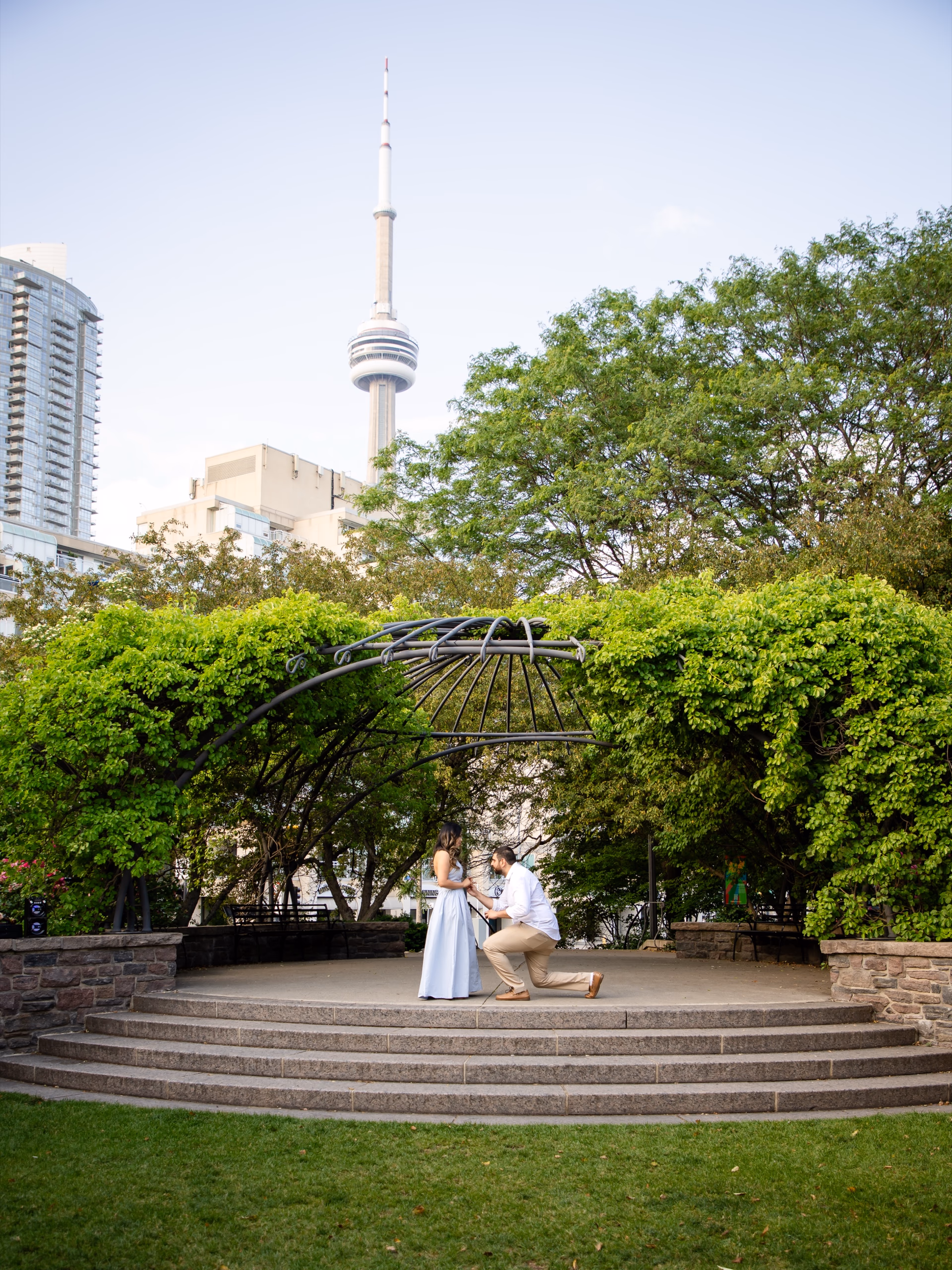A man on one knee proposing under the iron pavilion at the Toronto Music Garden with the CN Tower rising behind them, by AD Photography