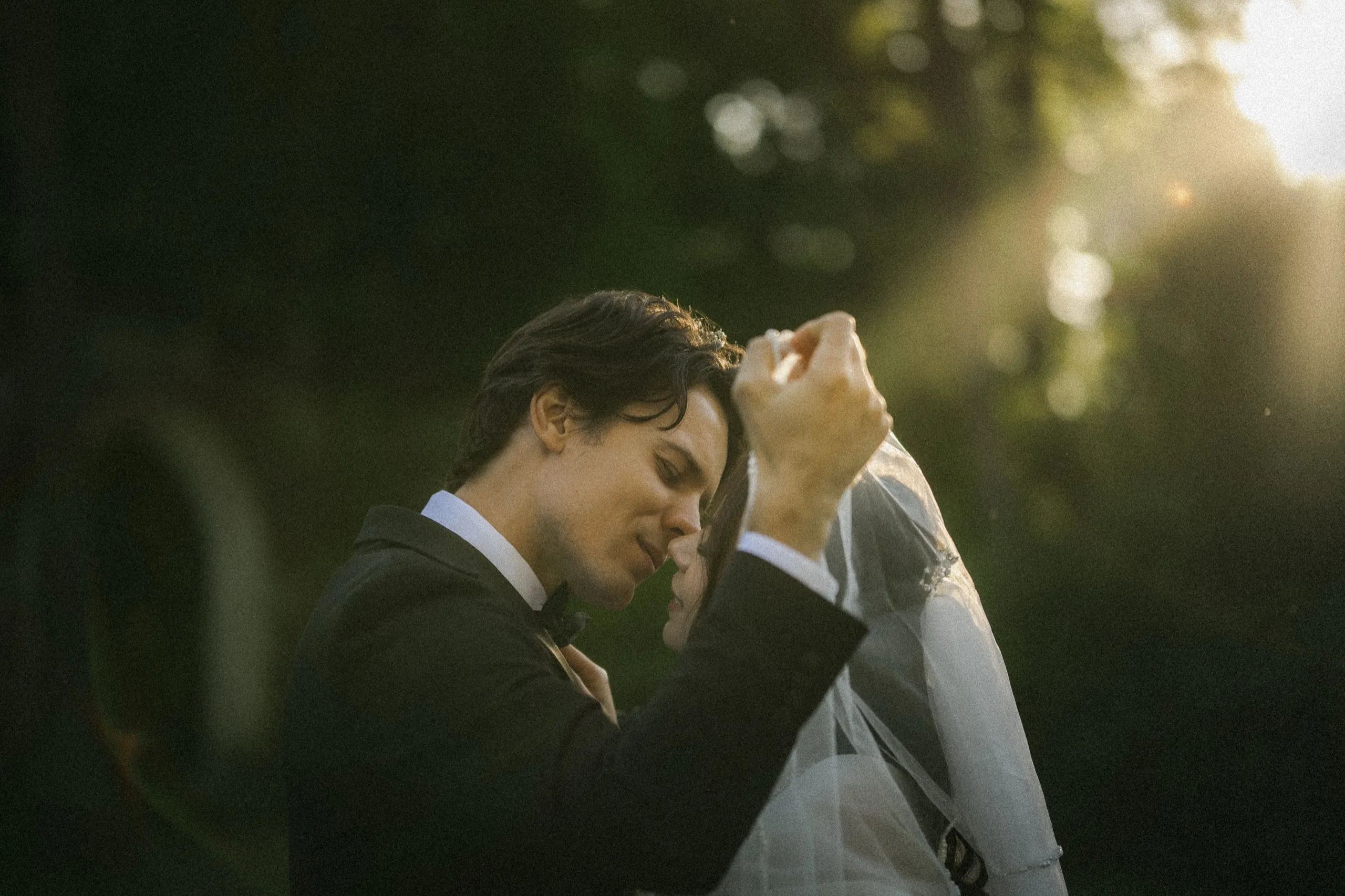Groom lifting the bride's blusher veil, nose to nose as sun rays fall through the trees at Guild Park & Gardens, by AD Photography
