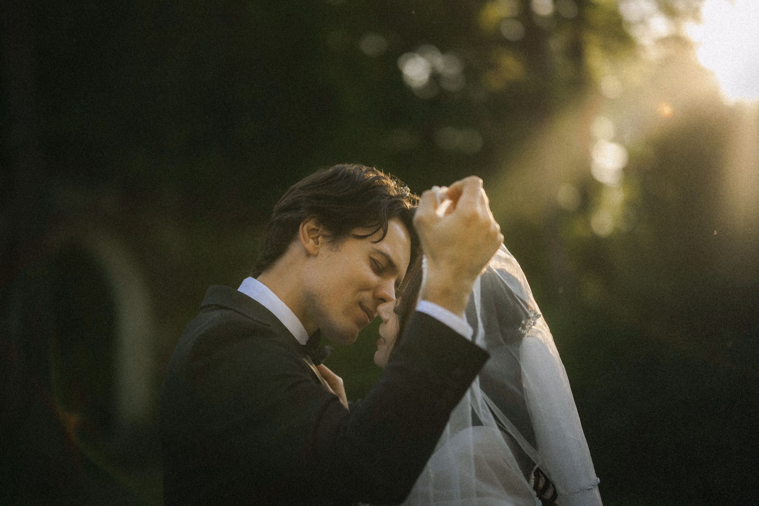 Groom lifting the bride's blusher veil, nose to nose as sun rays fall through the trees at Guild Park & Gardens, by AD Photography