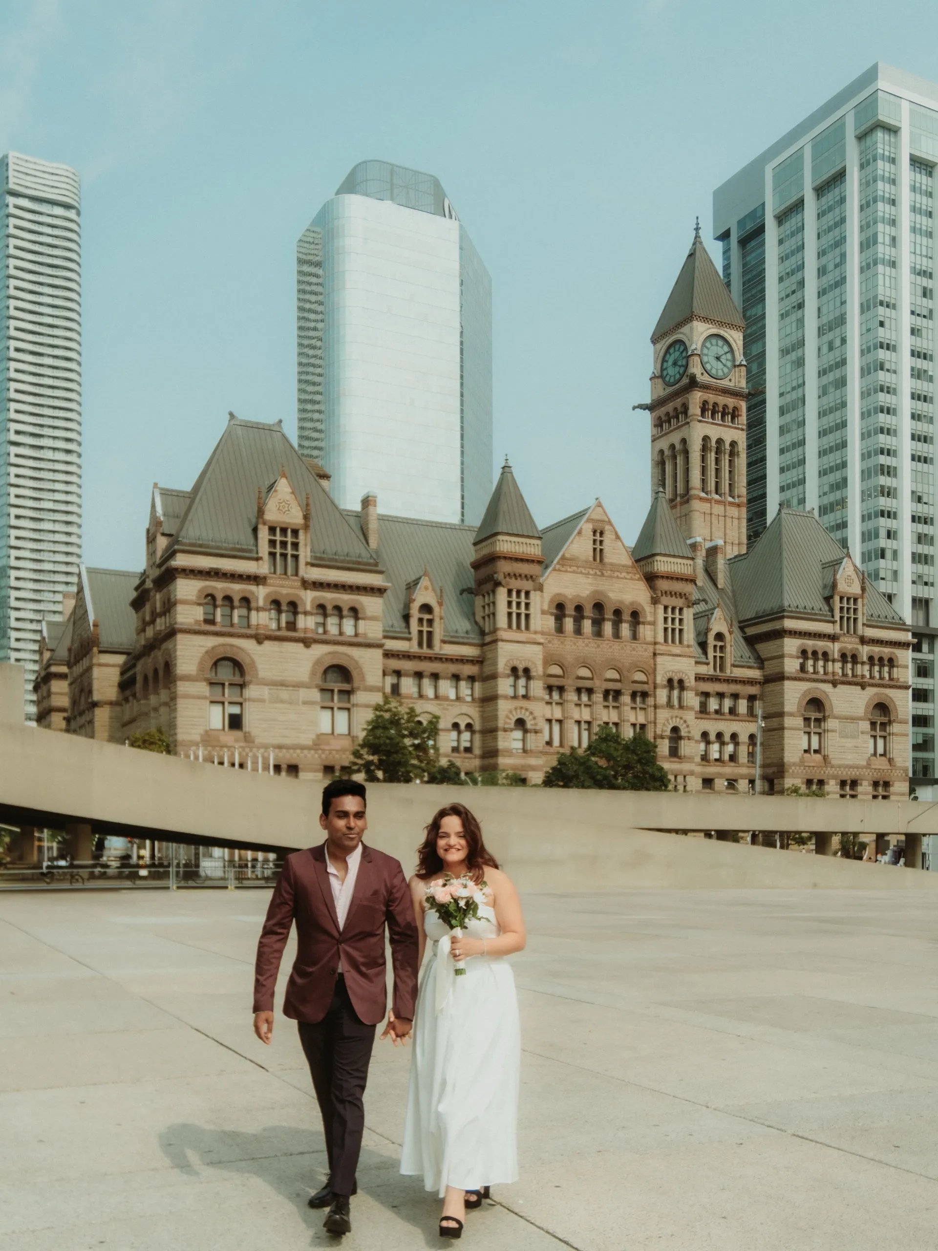A couple in wedding attire walk hand in hand across Nathan Phillips Square, the sandstone facade and clock tower of Toronto's Old City Hall rising behind them, by AD Photography