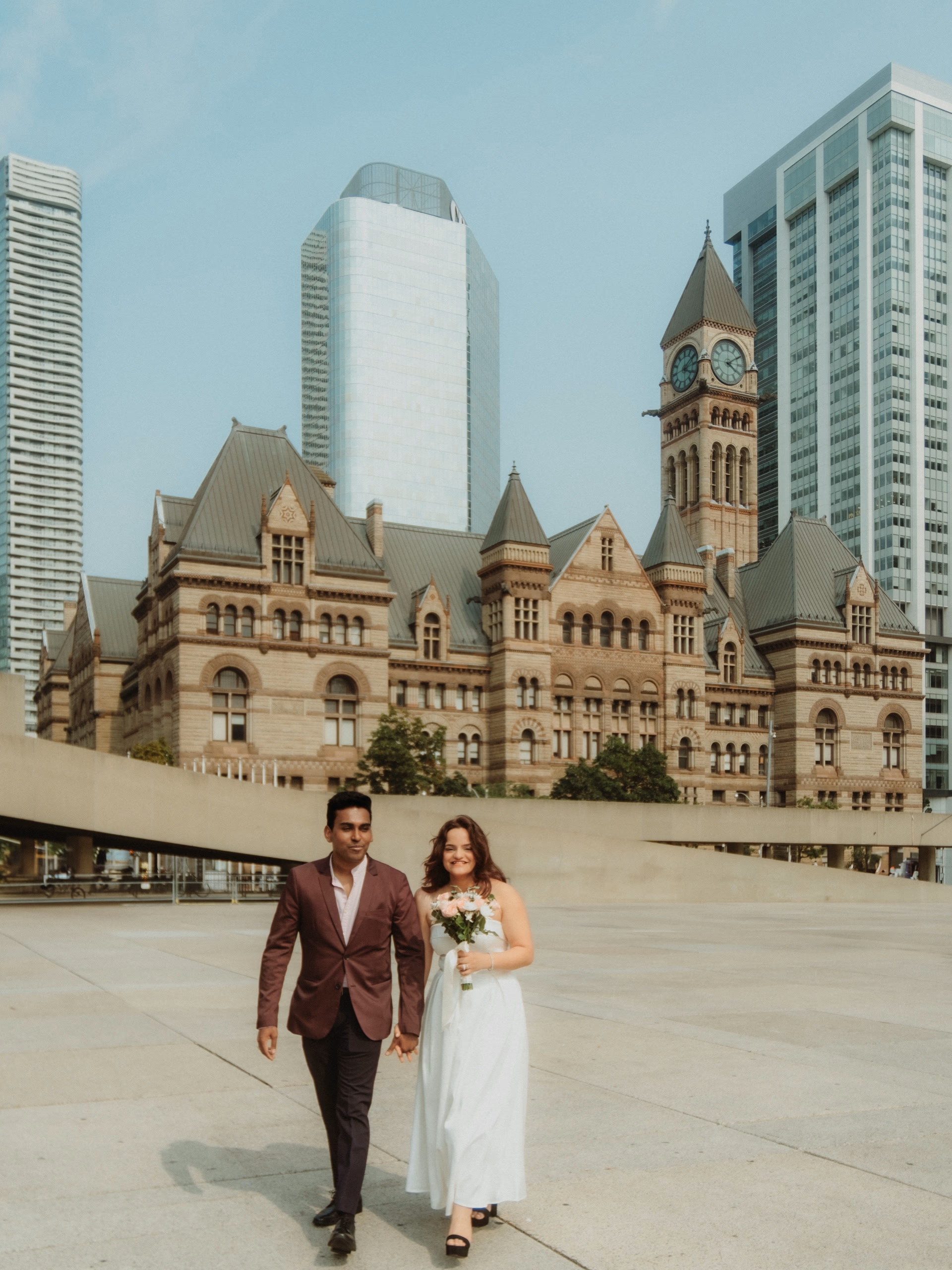 A couple in wedding attire walk hand in hand across Nathan Phillips Square, the sandstone facade and clock tower of Toronto's Old City Hall rising behind them, by AD Photography