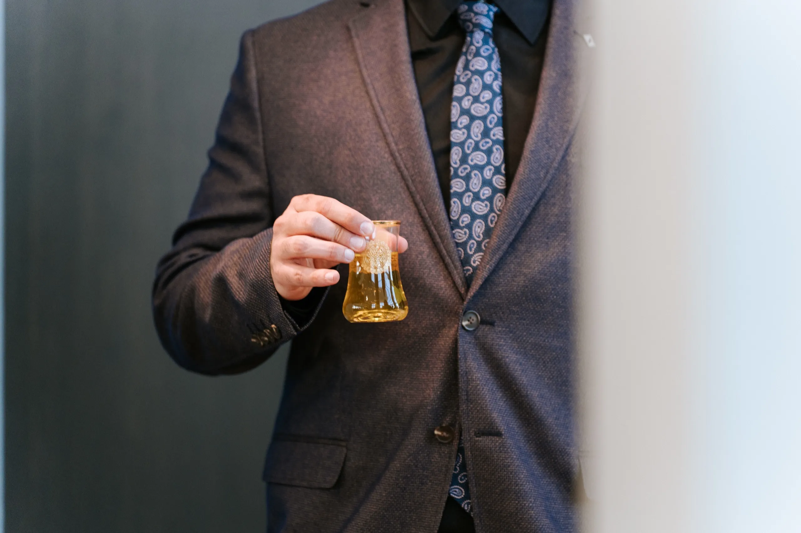 A guest in a brown tweed suit holds a small glass of tea during the celebration, by AD Photography