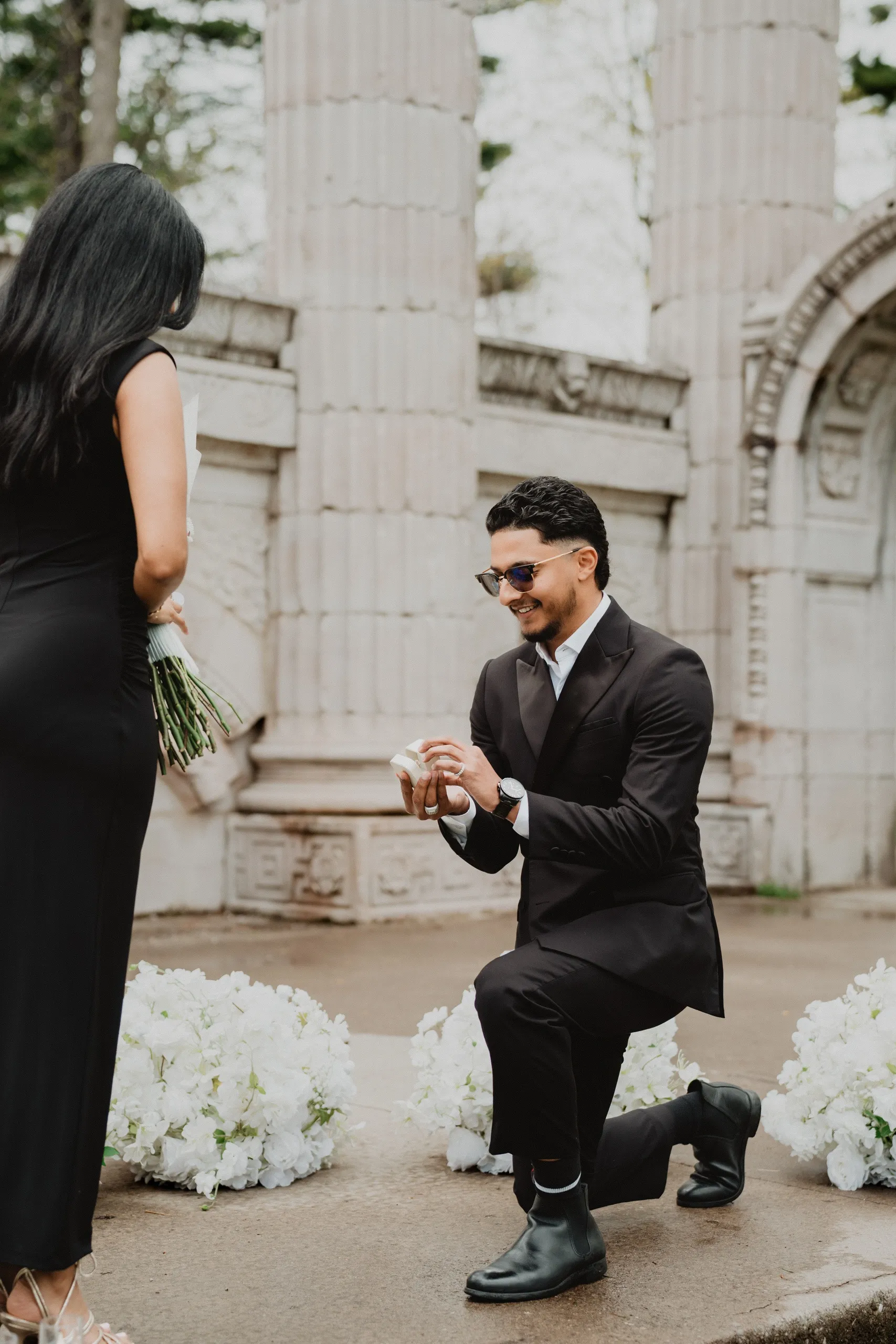 Raeed on one knee with the open ring box, the carved stone arch behind him in Scarborough, by AD Photography