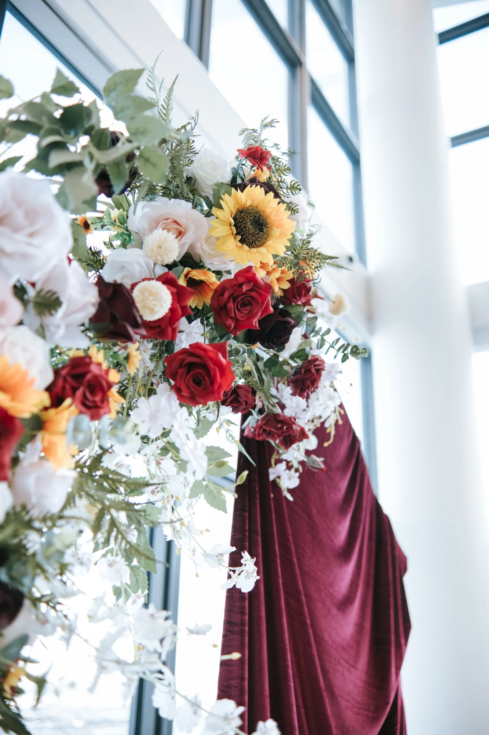 Sunflowers and red roses on the floral arch beside the burgundy drape and floor to ceiling windows, by AD Photography