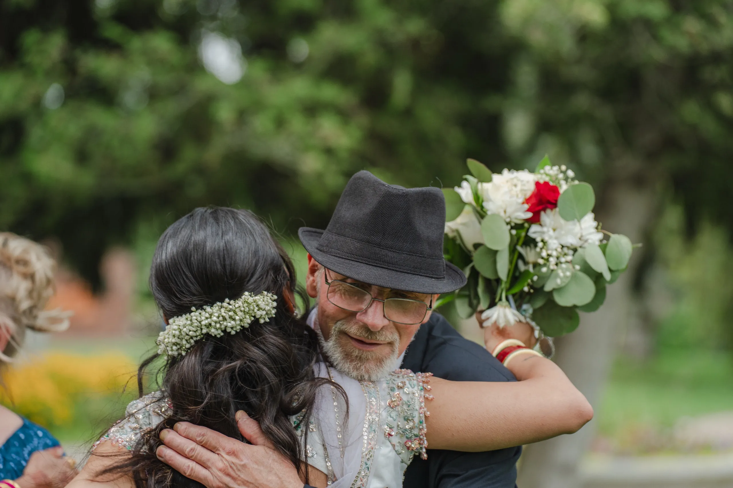 Nidhi shares a warm hug with a guest in a black fedora, bouquet held close., by AD Photography
