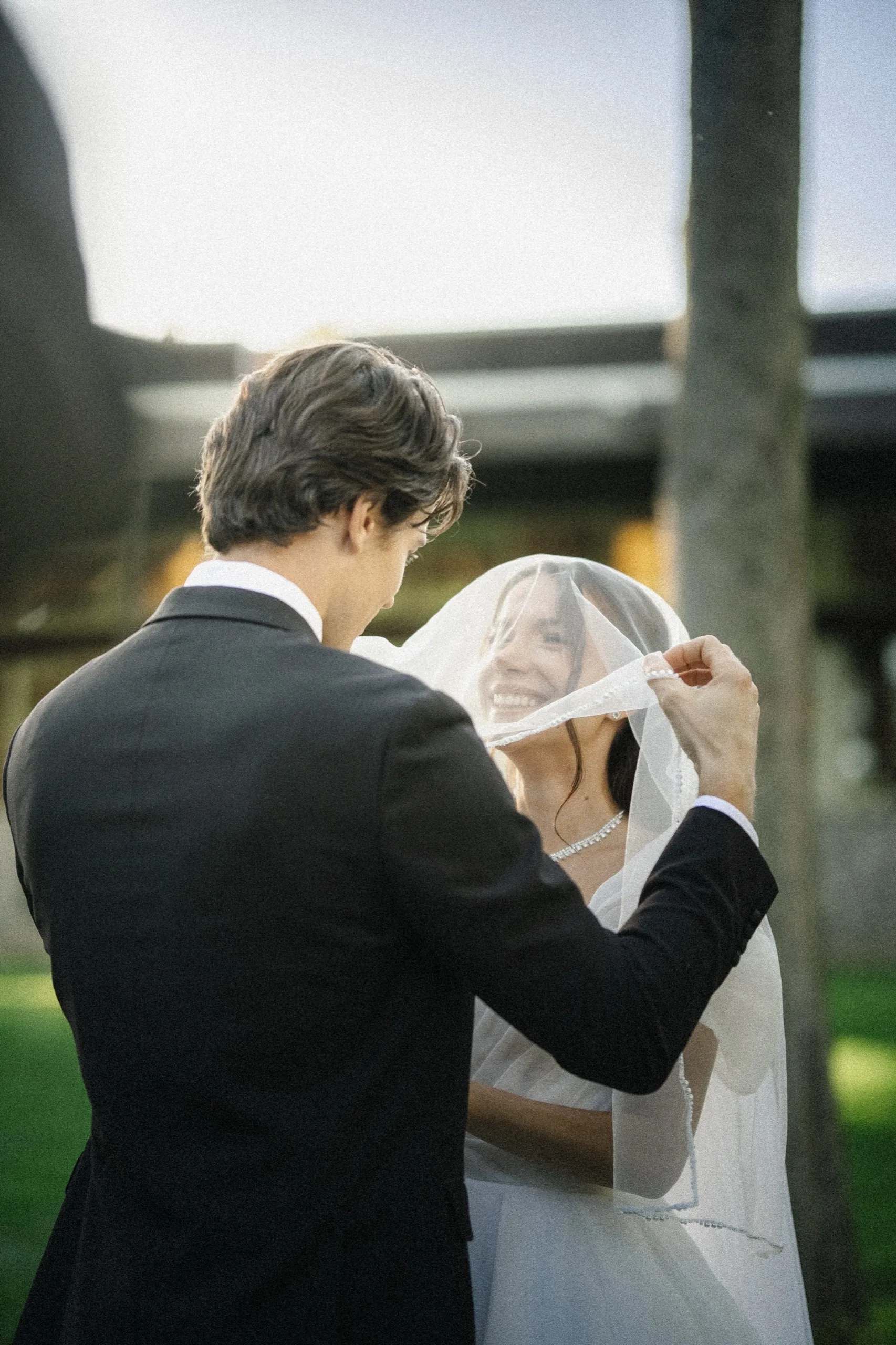 Bride laughing as the groom lifts her blusher veil in the evening light, by AD Photography
