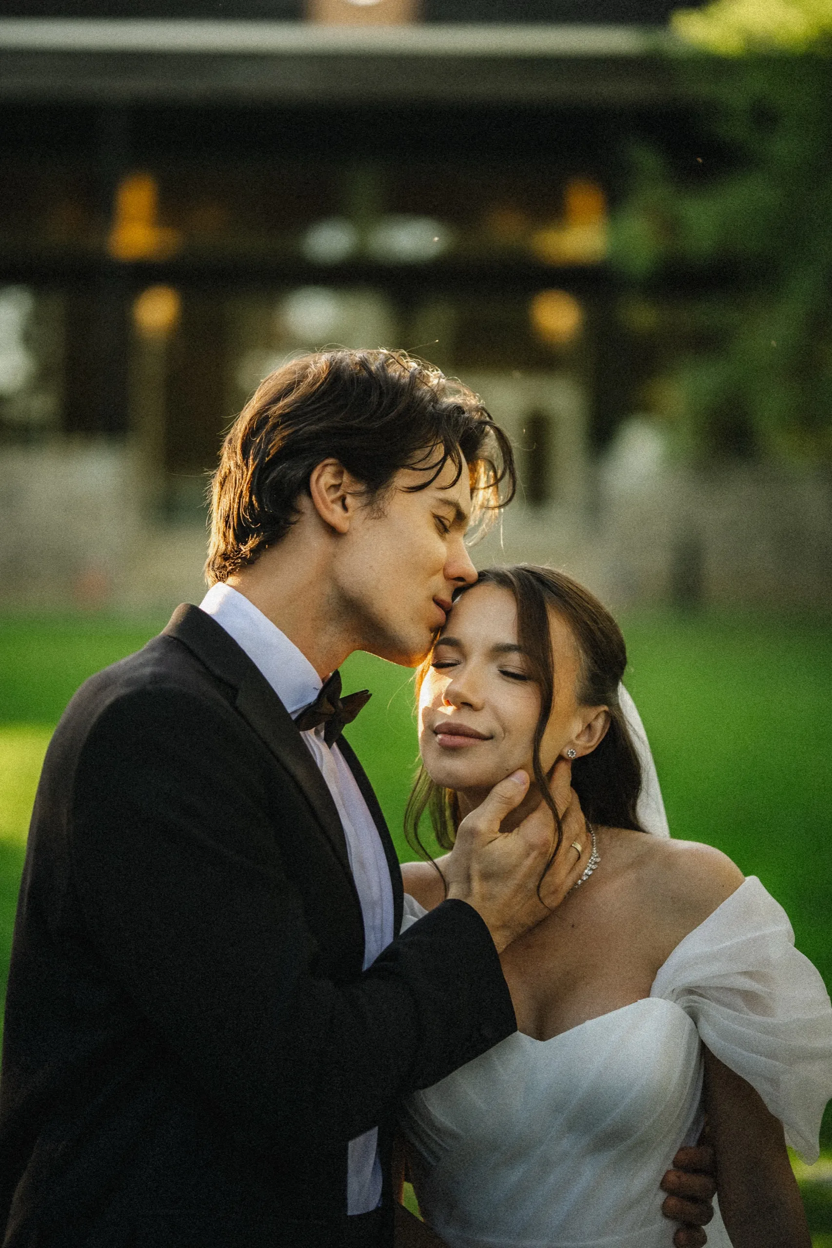 Groom kissing the bride's forehead, her eyes closed, warm side light across the lawn, by AD Photography