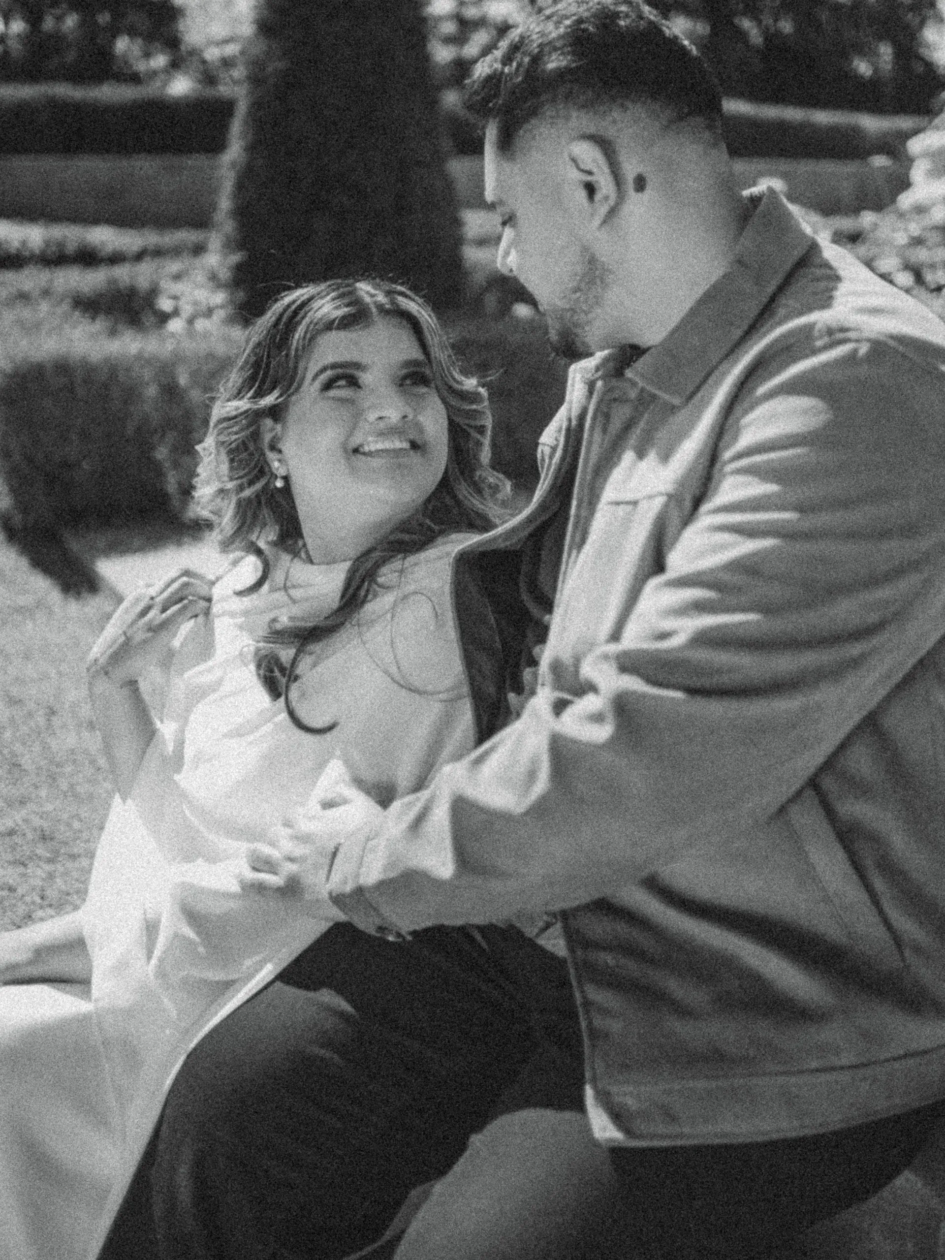 In black and white, Parneet smiles up at Maninder as they sit together in the Formal Garden, by AD Photography