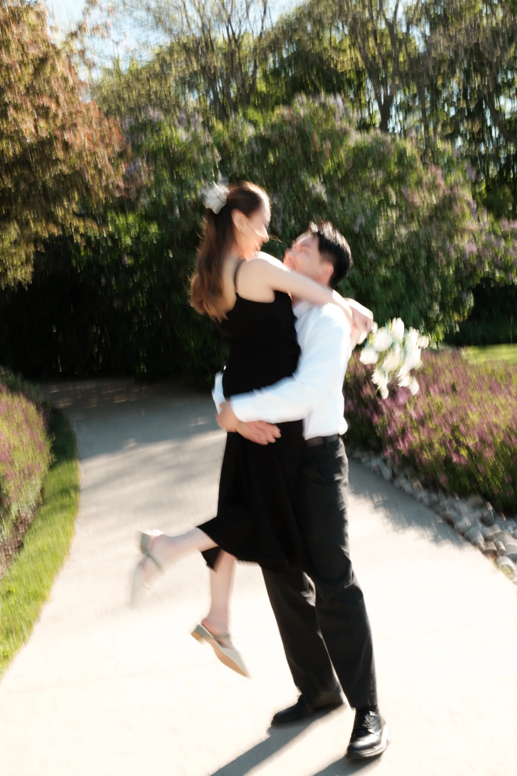 Motion blur of Nelson lifting Josie off her feet in a spin among the garden greenery, by AD Photography