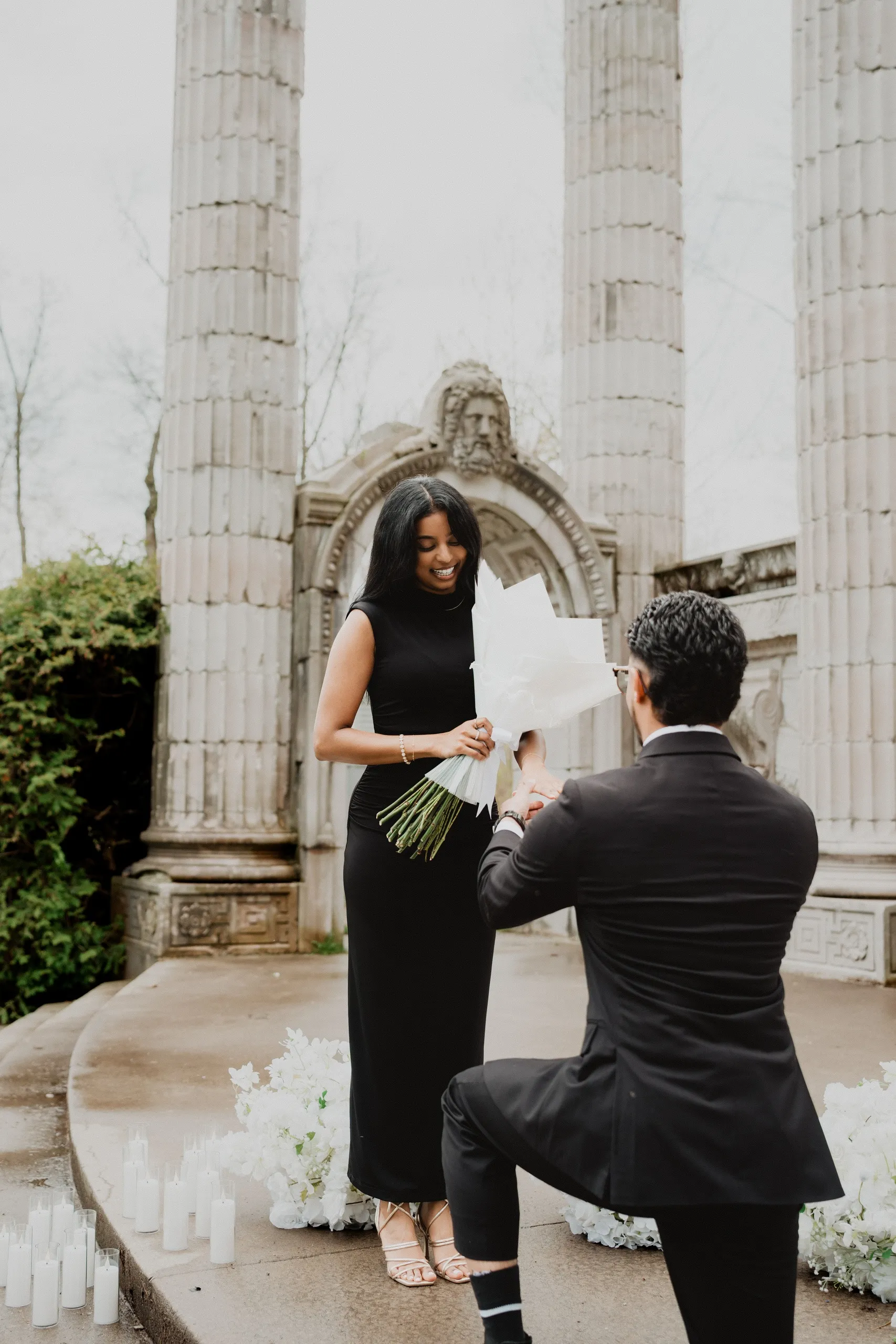 Raeed kneeling to hand Lavina the bouquet as she laughs under the colonnade, by AD Photography