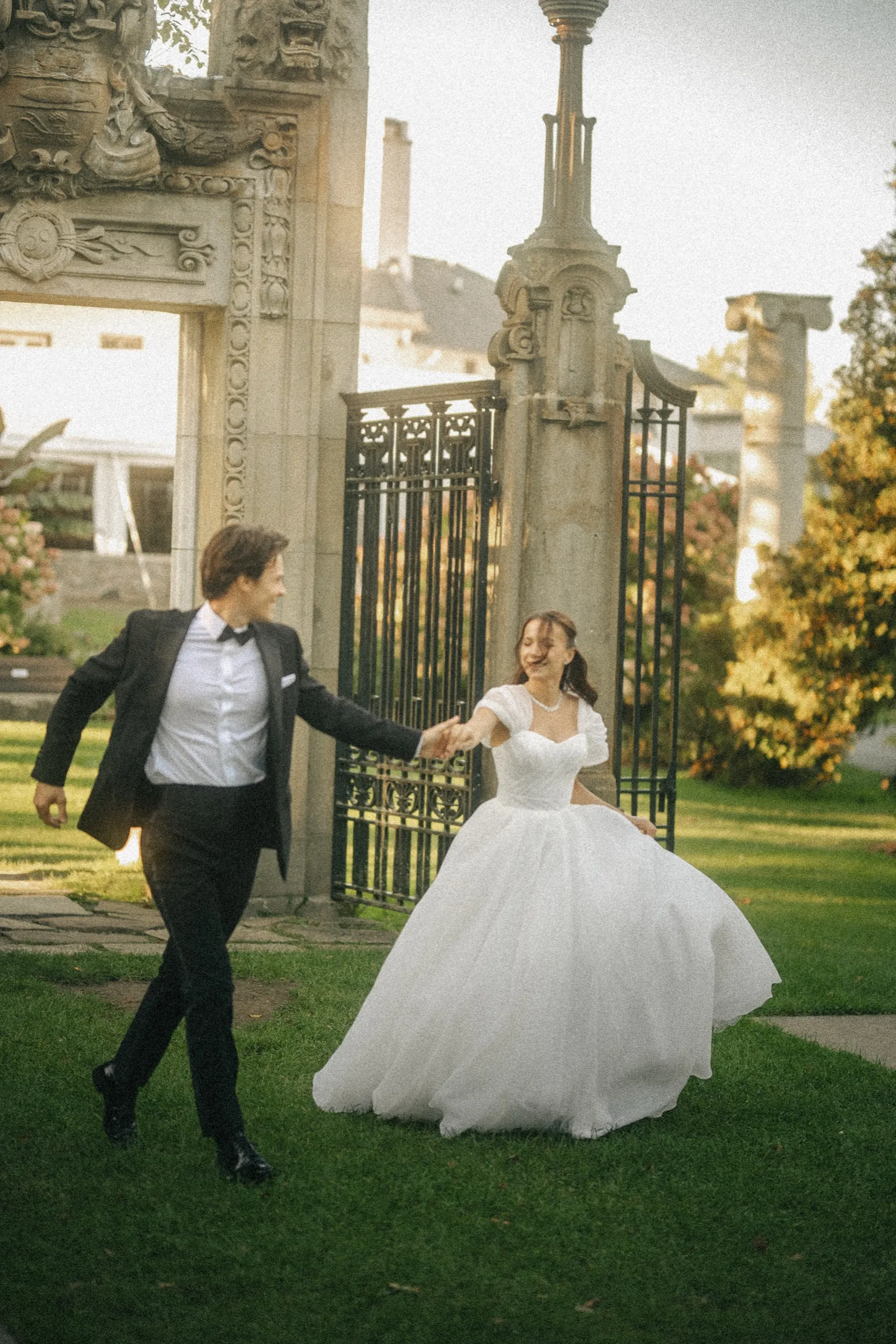Bride and groom running hand in hand past the ornate iron gate and carved stone gateway, by AD Photography