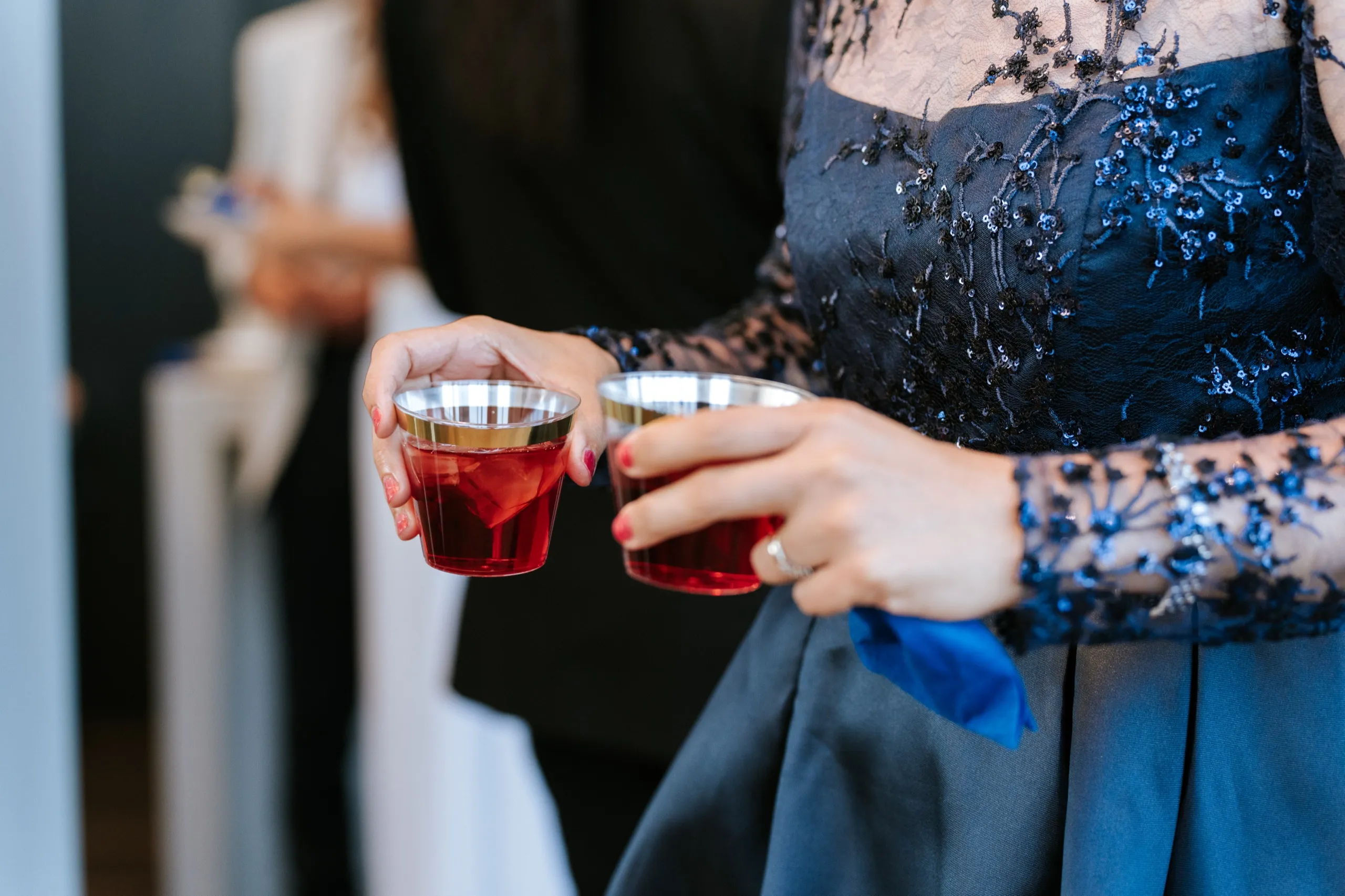 A guest in a navy beaded gown carries two cups of red tea through the room, by AD Photography