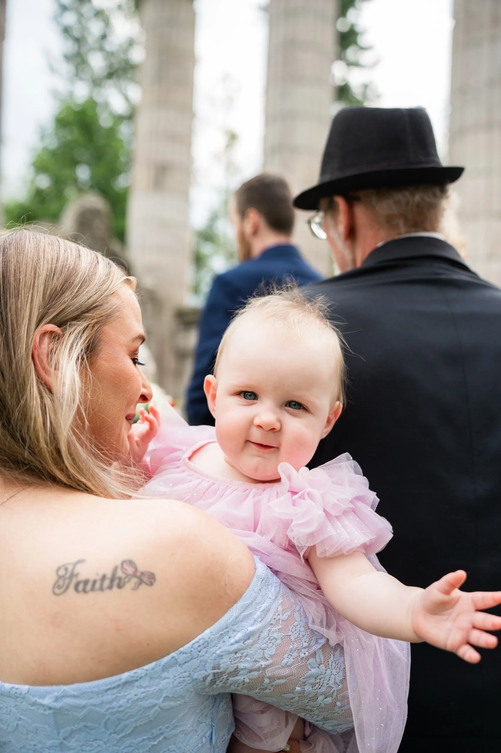 A guest holds a baby in pink tulle as family gathers beneath the columns nearby., by AD Photography