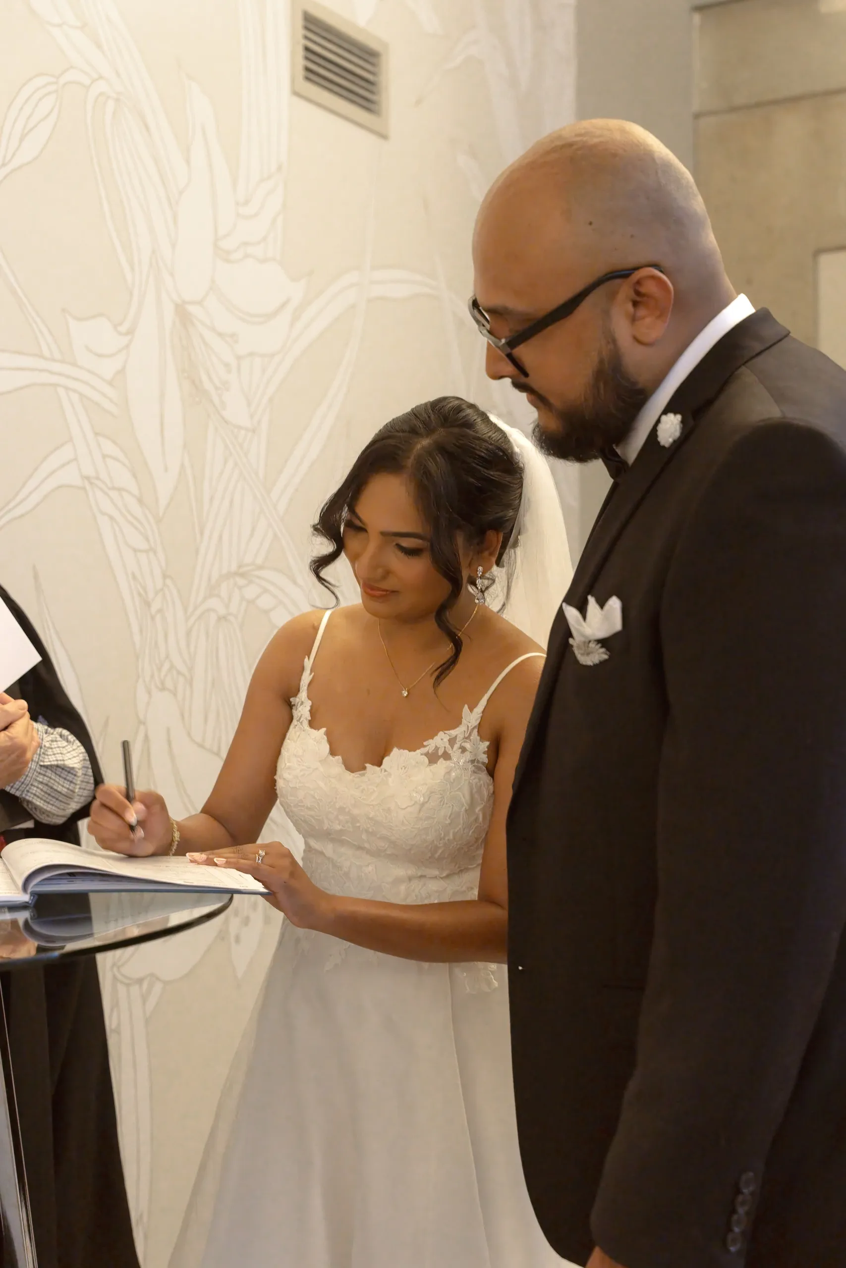Bride signs the marriage register while the officiant looks on, by AD Photography