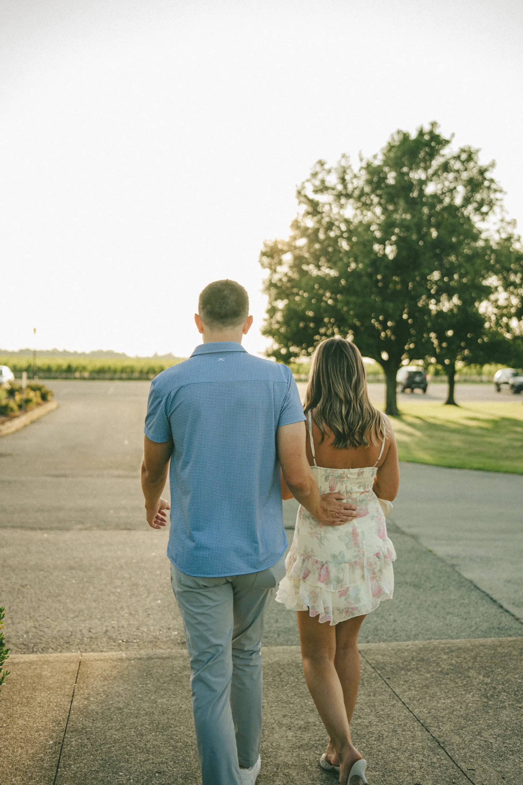 Donald's hand low on Ashley's back as they walk toward the vineyard in blown out afternoon light, by AD Photography