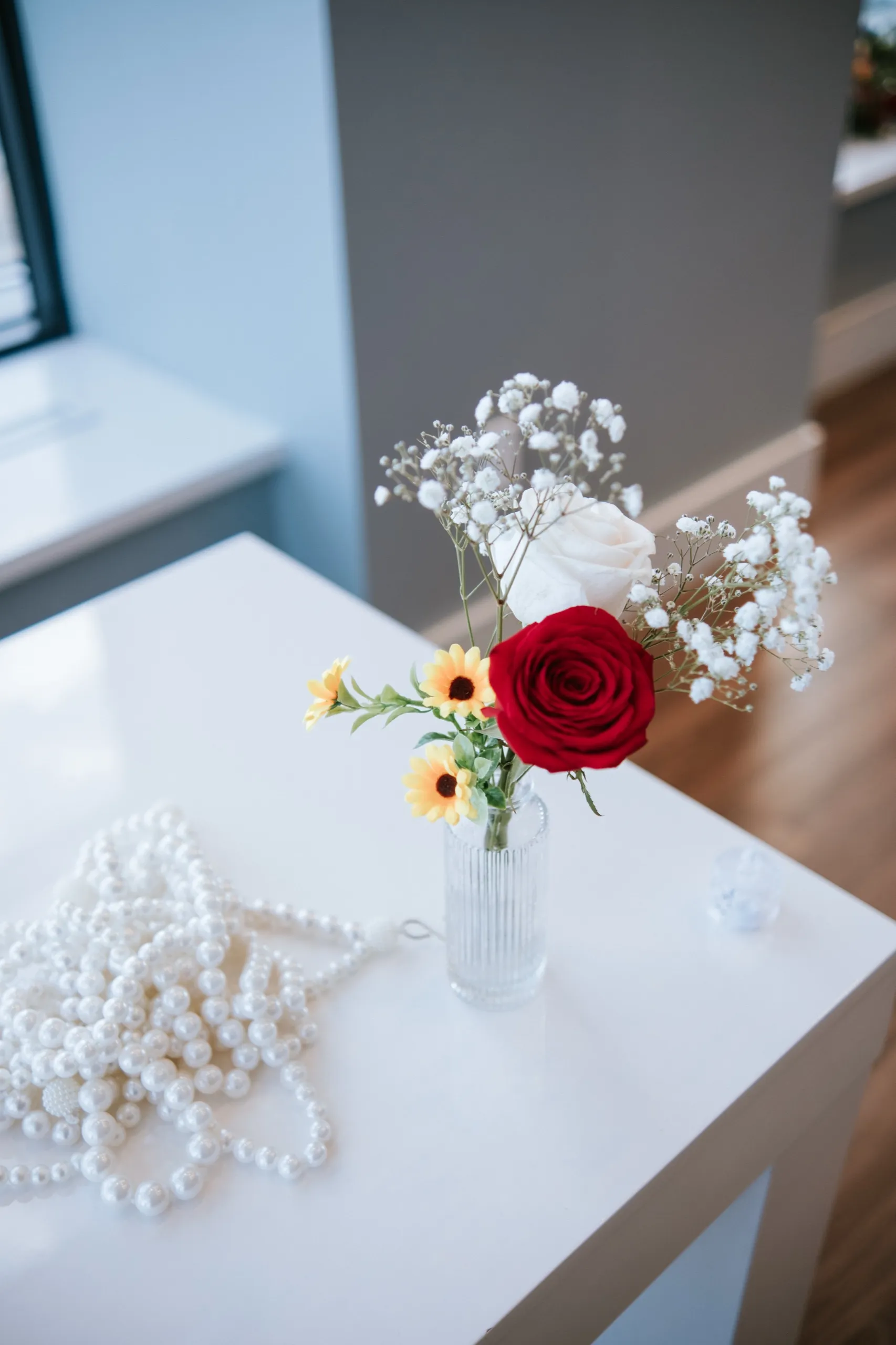 Red rose, sunflowers and baby's breath in a small vase beside strands of pearls, by AD Photography