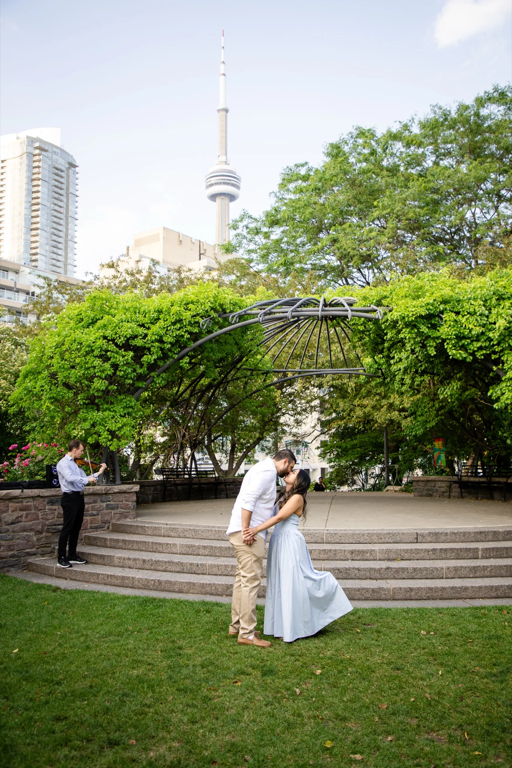 Newly engaged couple kissing on the Toronto Music Garden amphitheatre stage with the CN Tower behind them, by AD Photography