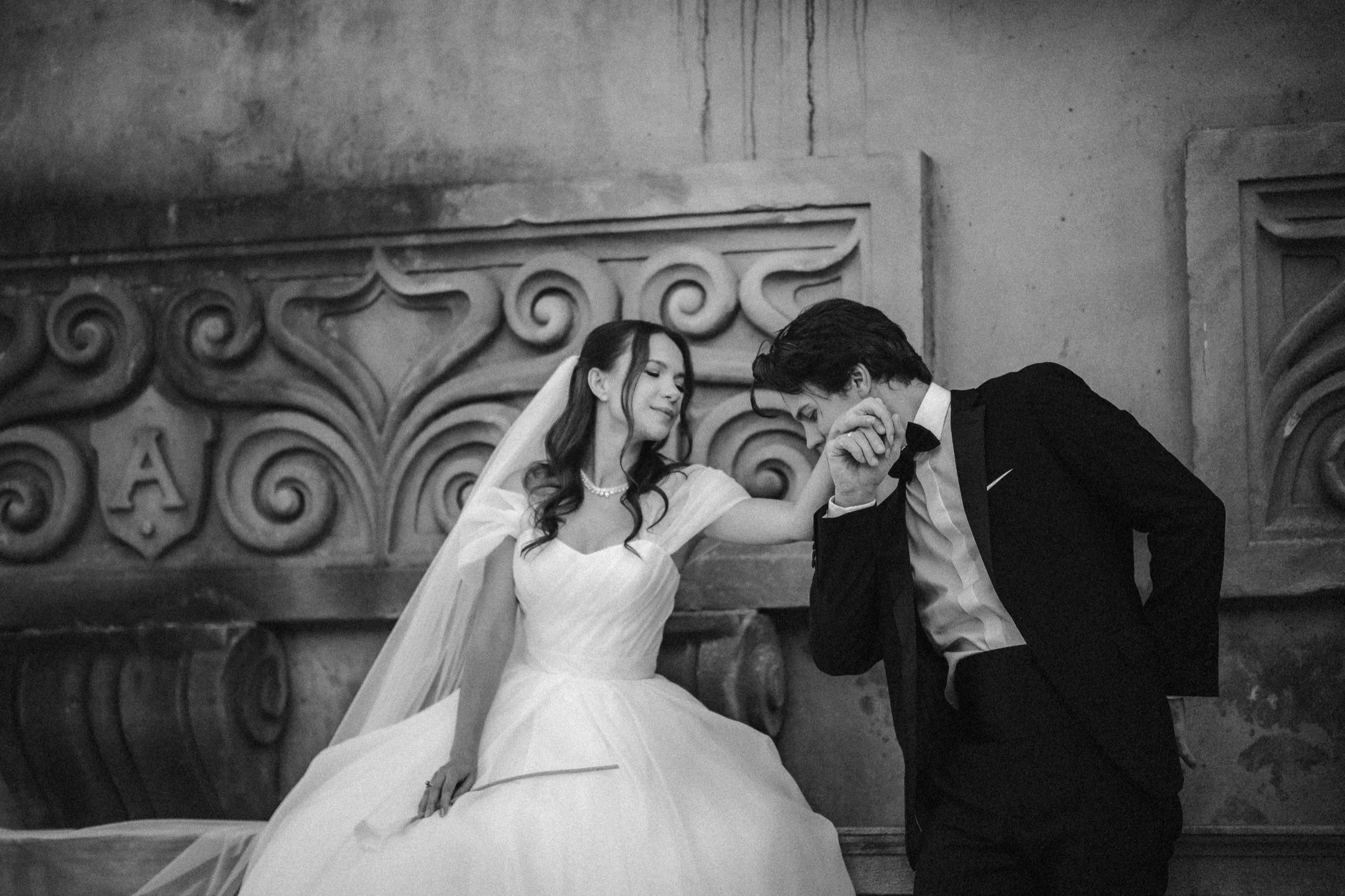 Black and white of the groom kissing the bride's hand against a carved stone scroll relief, by AD Photography