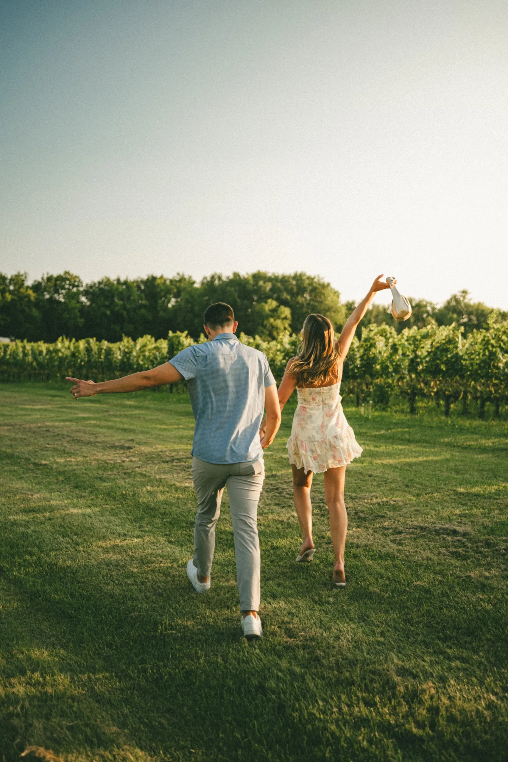 Ashley raising the champagne bottle overhead as the two of them walk out into the vines, by AD Photography