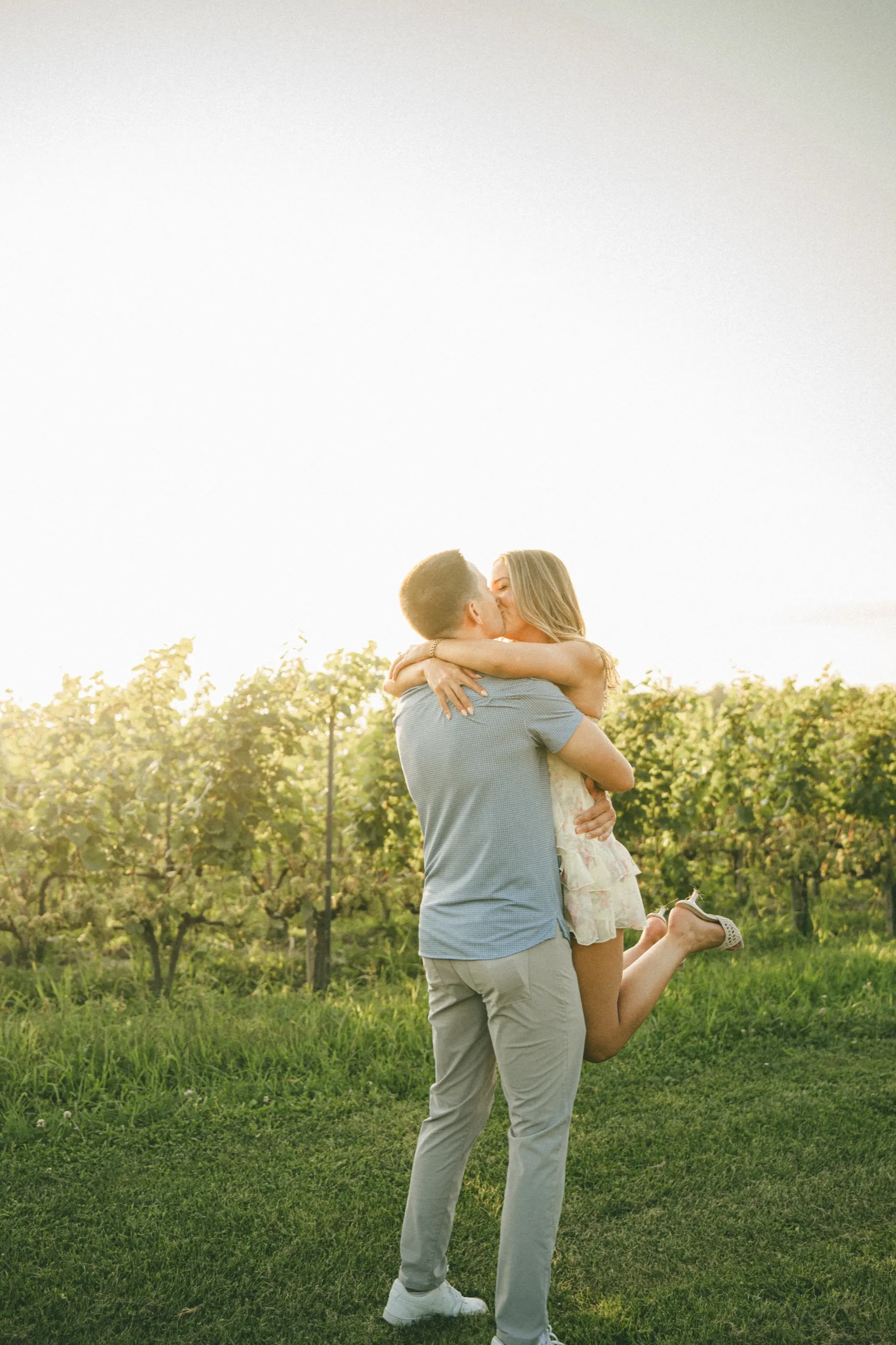 Donald lifting Ashley off her feet for a kiss in the vine rows, backlit by the low sun, by AD Photography