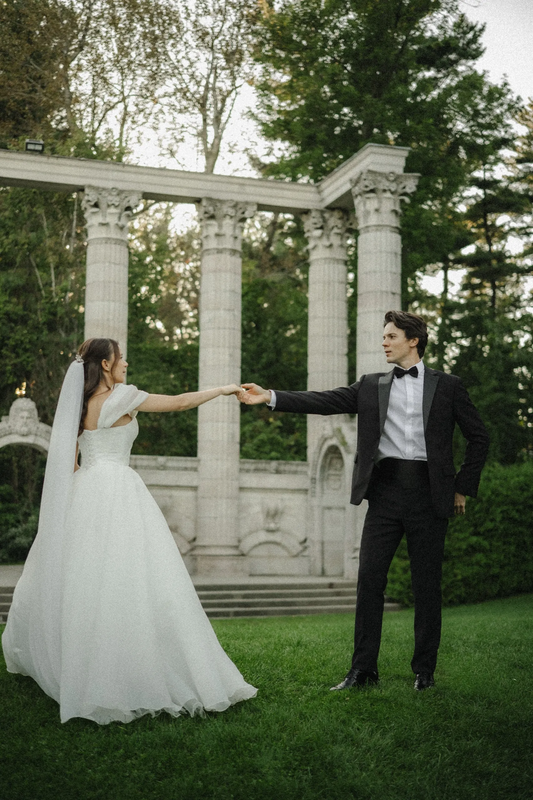 Bride and groom at arm's length about to twirl in front of the Guild Park colonnade in soft evening light, by AD Photography