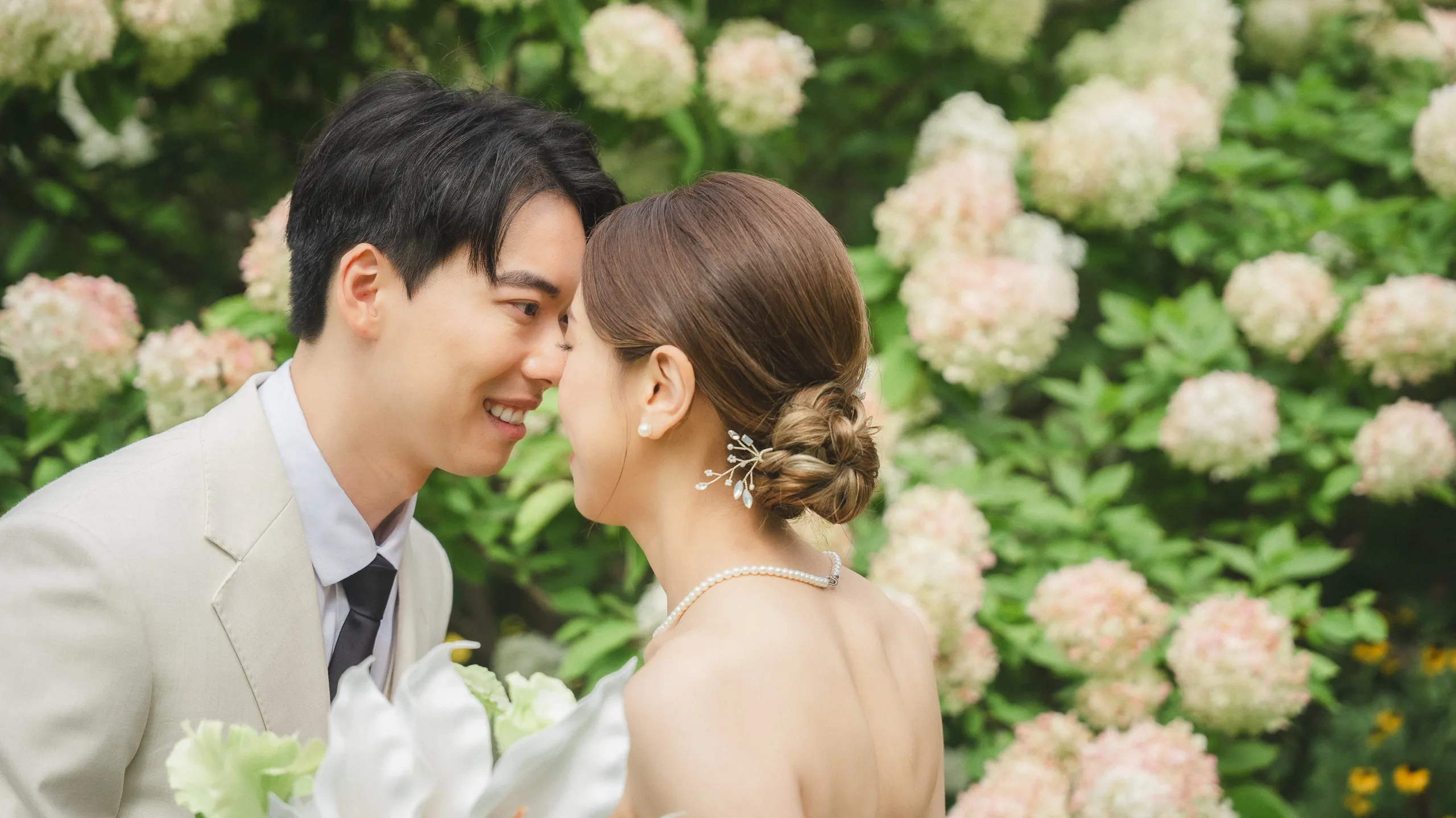 Josie and Nelson smiling forehead to forehead against a wall of blush hydrangeas, by AD Photography