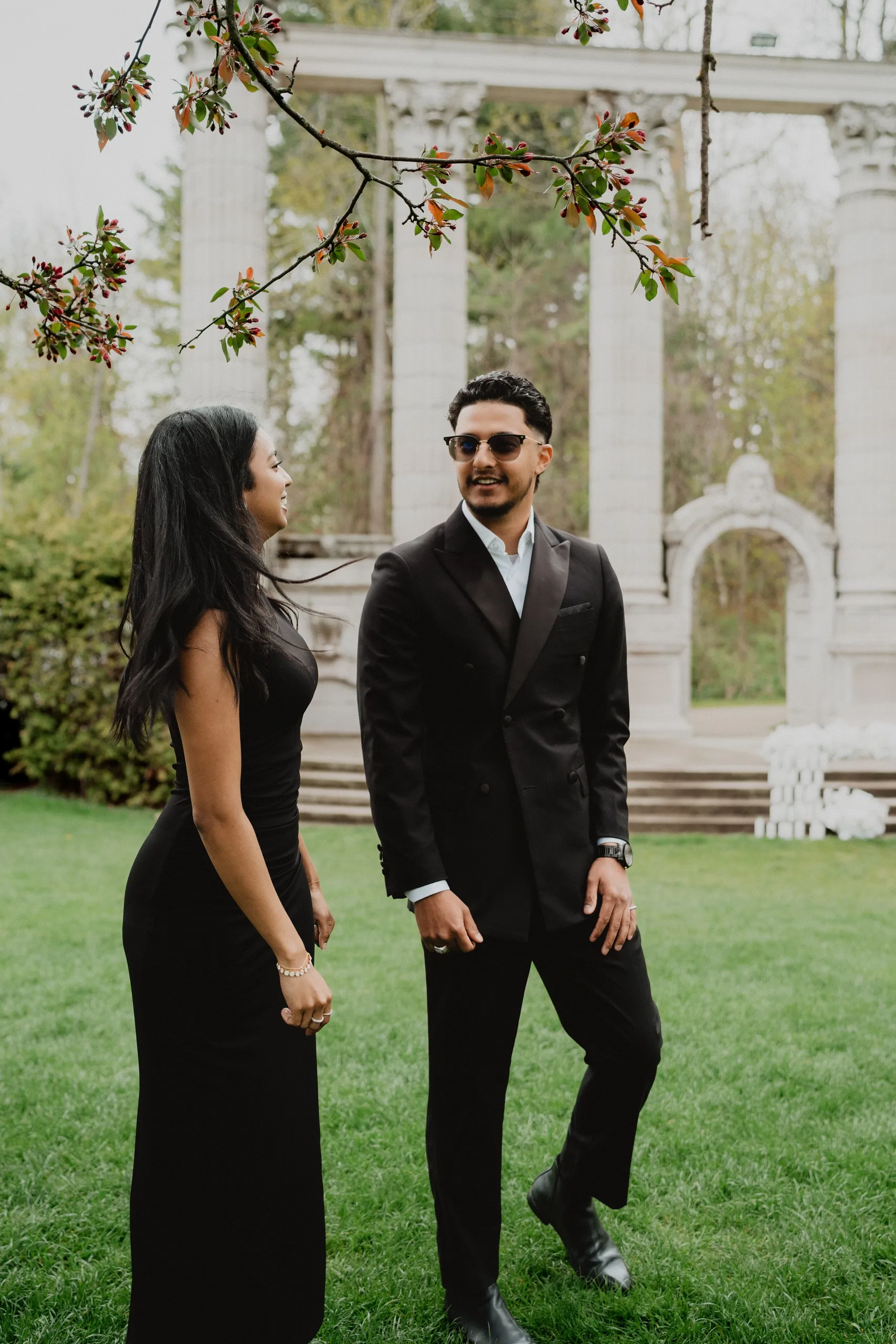 Lavina laughing at Raeed under a crabapple branch in bud beside the colonnade, by AD Photography