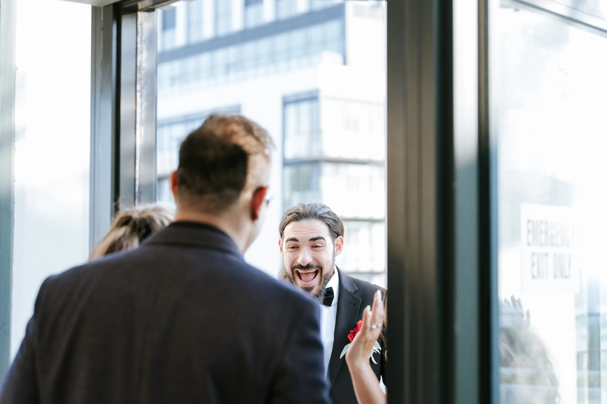Moe throws his hands up laughing as another guest arrives, caught through the glass doors, by AD Photography