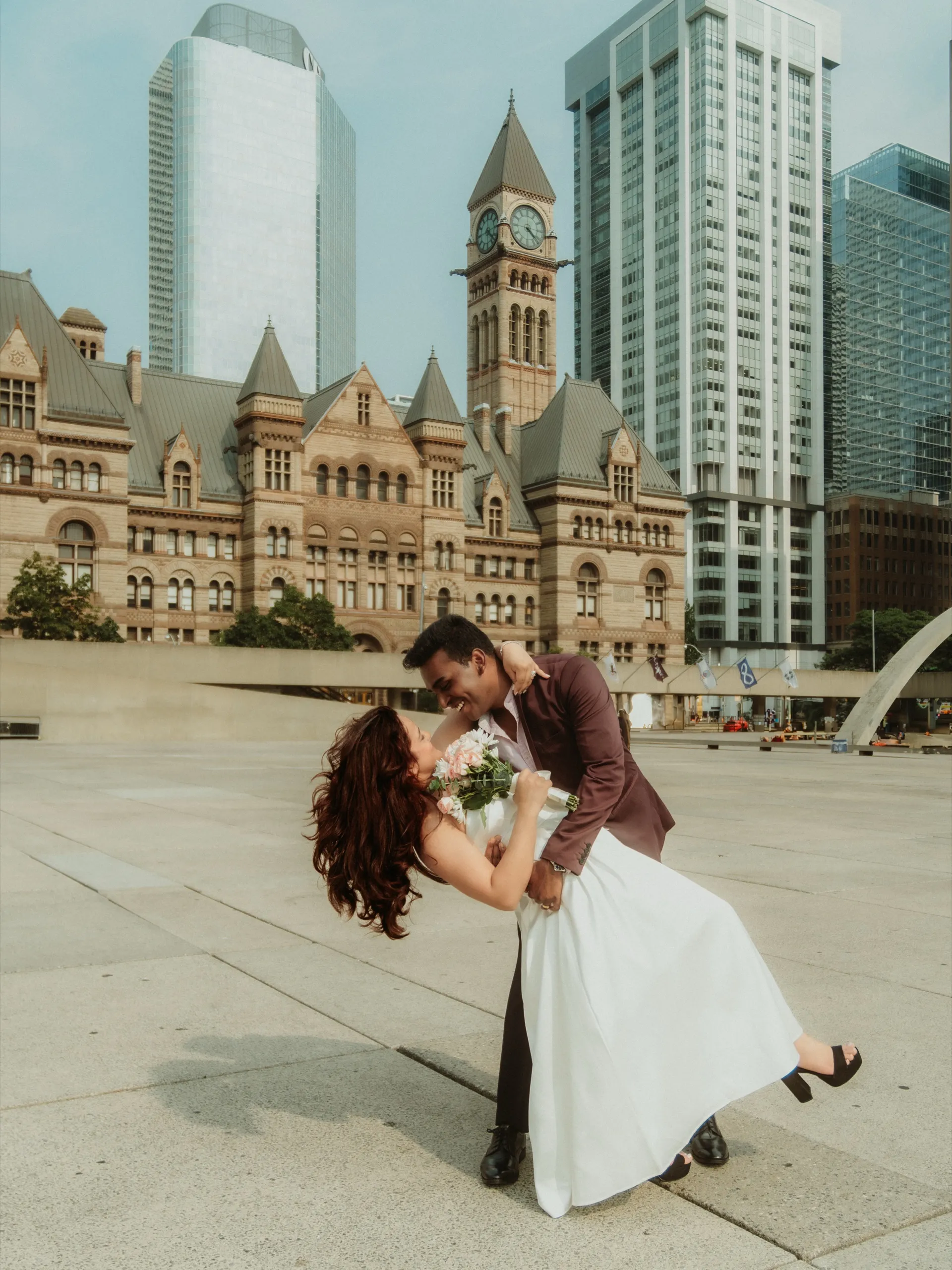 Anushka and Anthony, a dip kiss in Nathan Phillips Square after their Toronto City Hall elopement, Old City Hall clock tower behind them