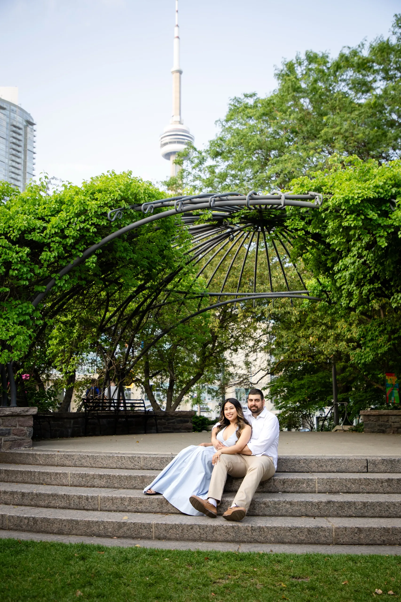 The newly engaged couple sitting on the granite amphitheatre steps at the Toronto Music Garden below the CN Tower, by AD Photography