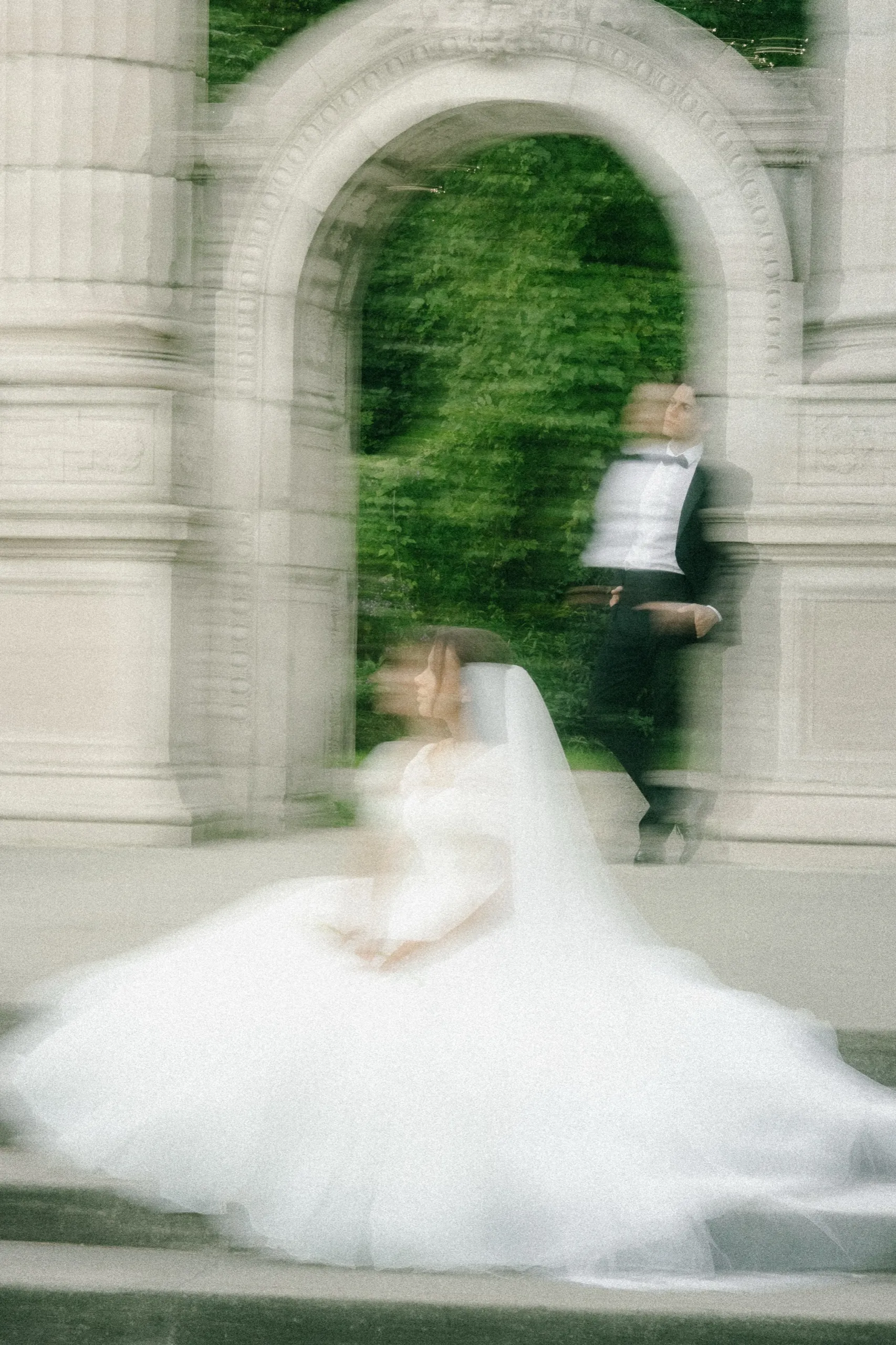 Slow shutter ghosting of the bride seated on the stone steps and the groom standing in the carved archway at Guild Park, by AD Photography