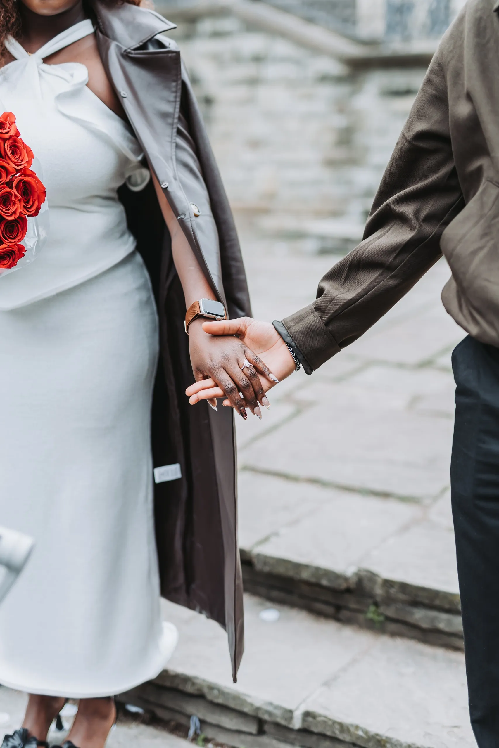 Their joined hands and her engagement ring, red roses tucked against her white dress, by AD Photography