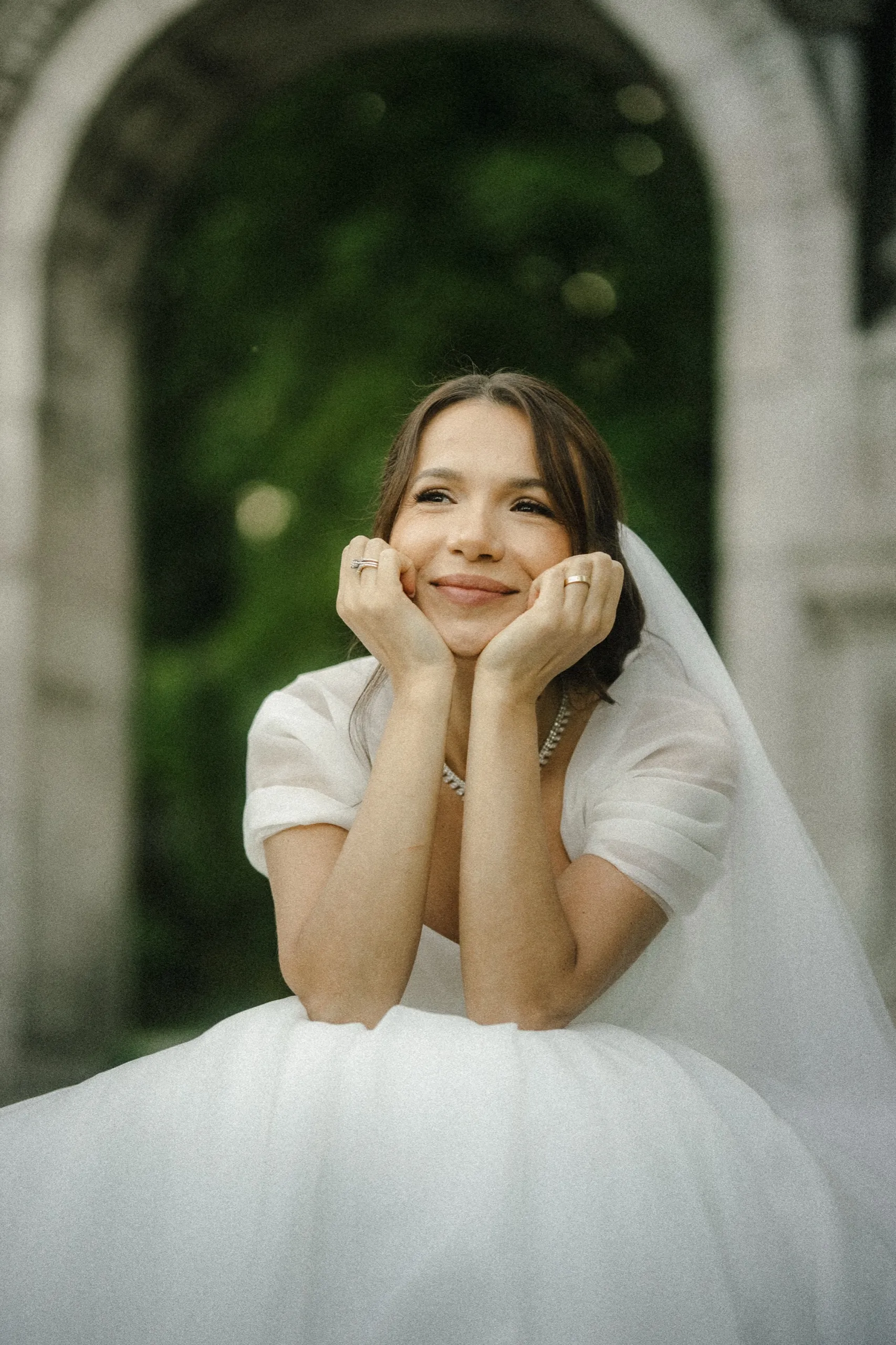 Bride resting her chin on her hands and smiling, the stone archway soft behind her, by AD Photography