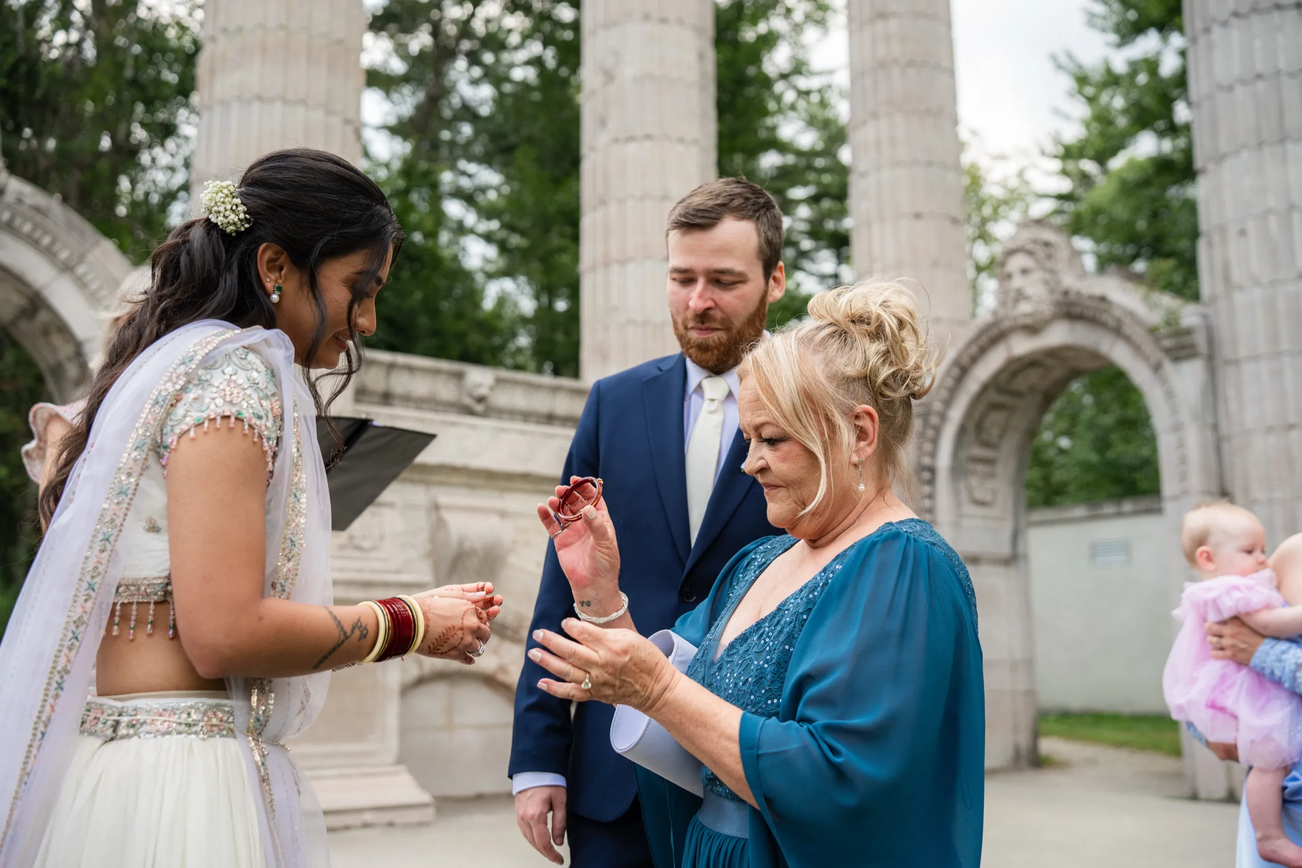 Nidhi's mother in law presents the wedding bands while Derek watches, a baby fussing nearby., by AD Photography