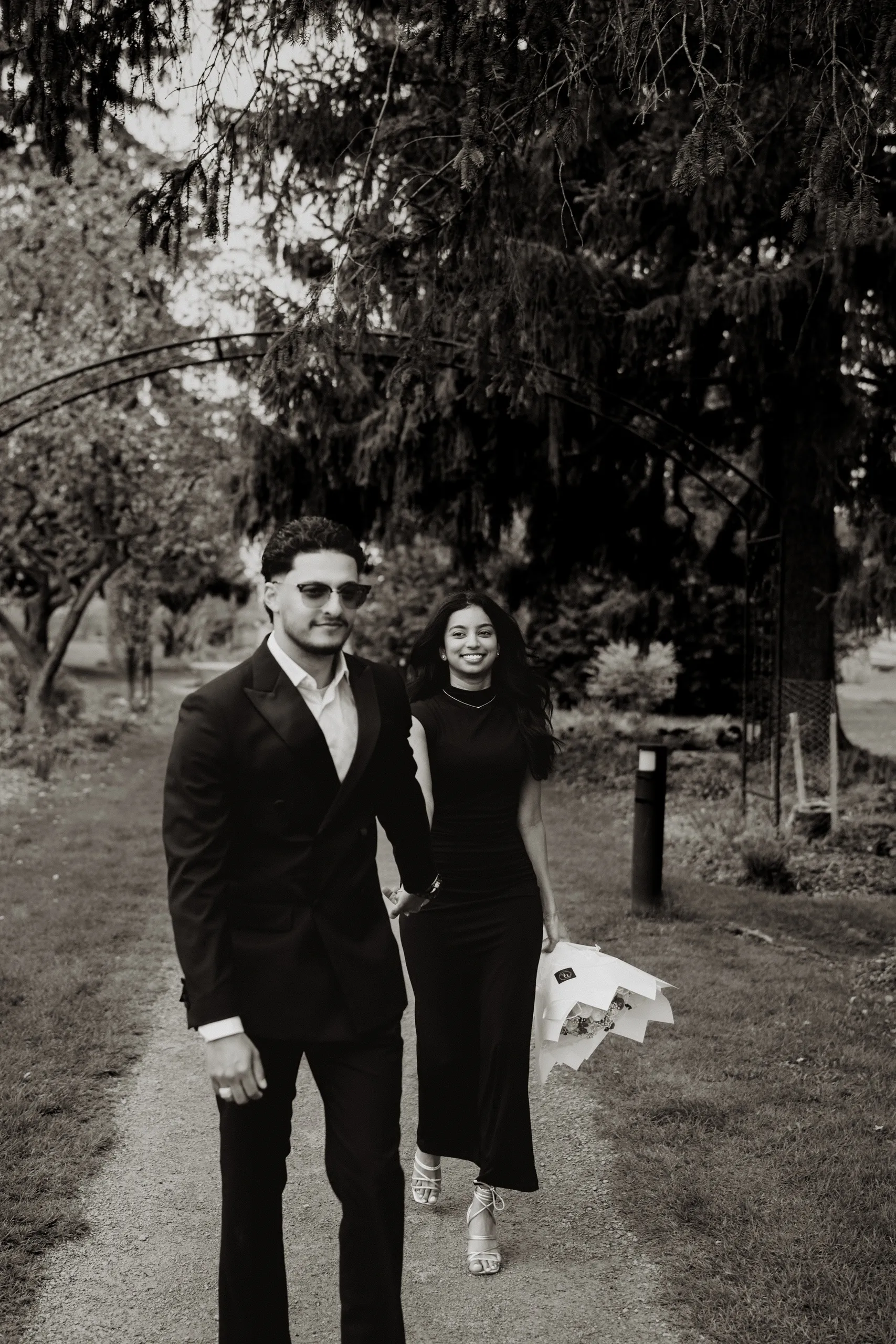 A black and white frame of the couple walking a garden path under old cedars in Scarborough, by AD Photography