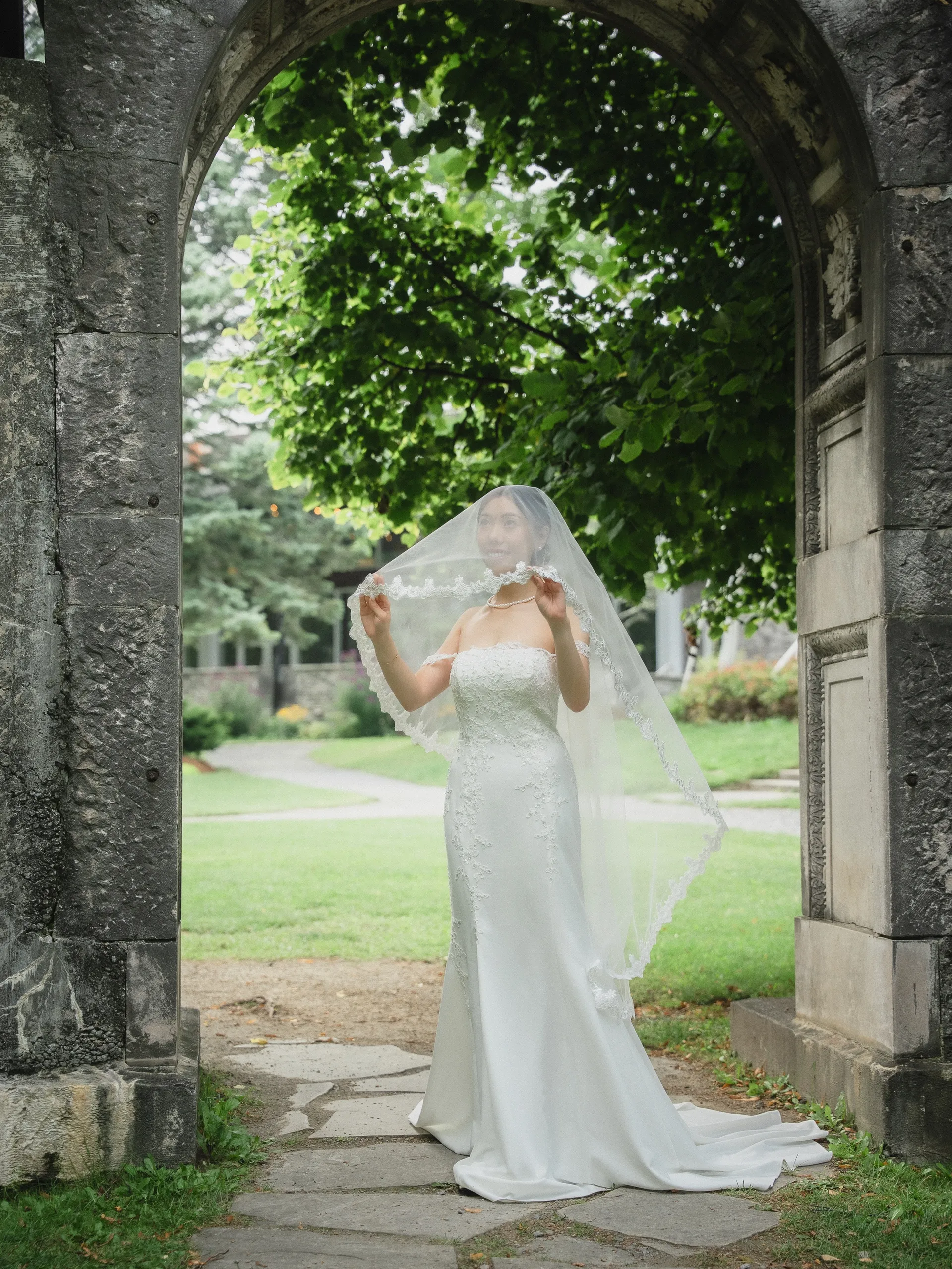 Josie holding her lace veil open wide under the stone arch at Guild Park, by AD Photography