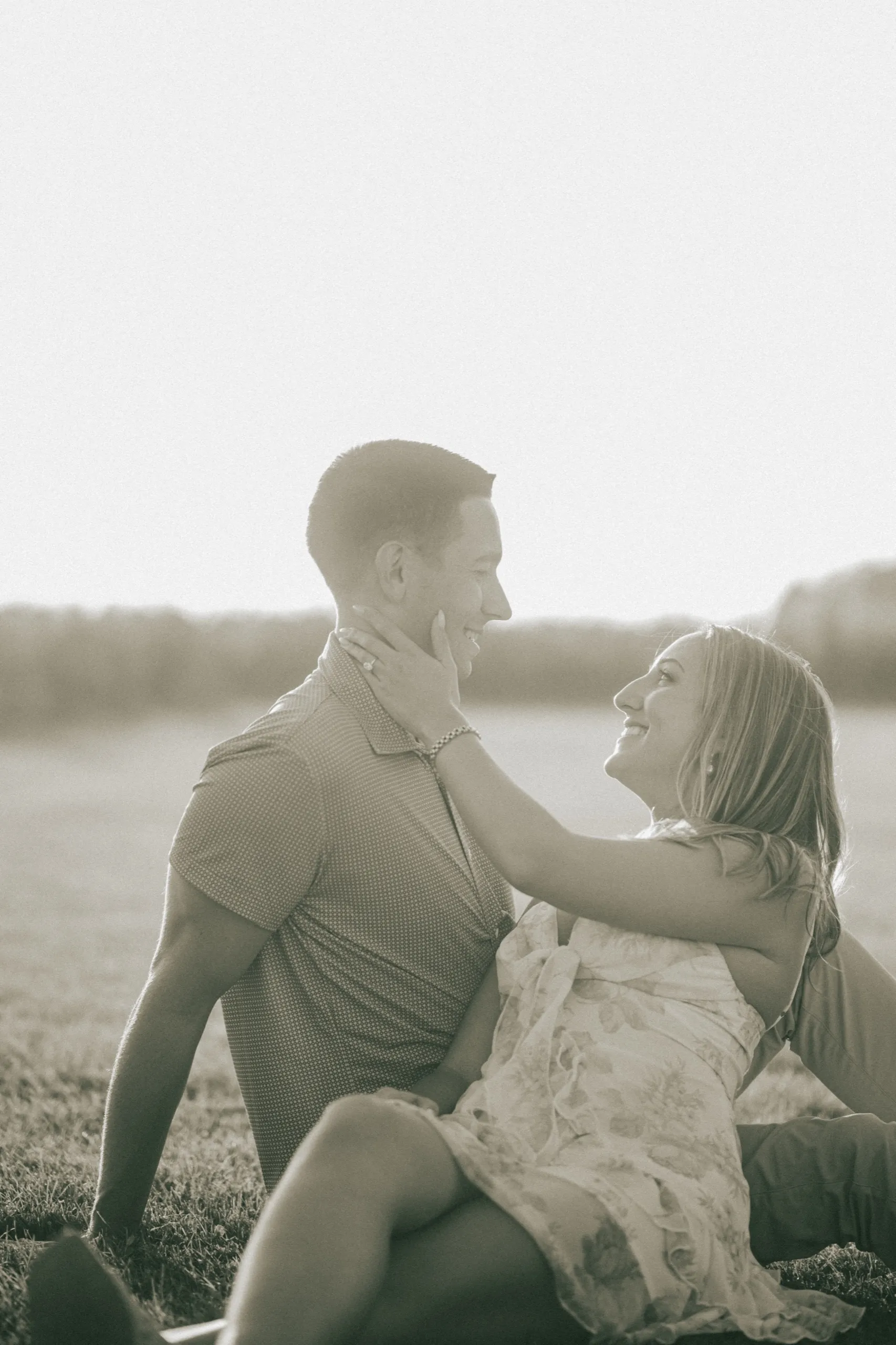 A black and white frame of Ashley sitting into Donald on the grass, her hand at his jaw, by AD Photography