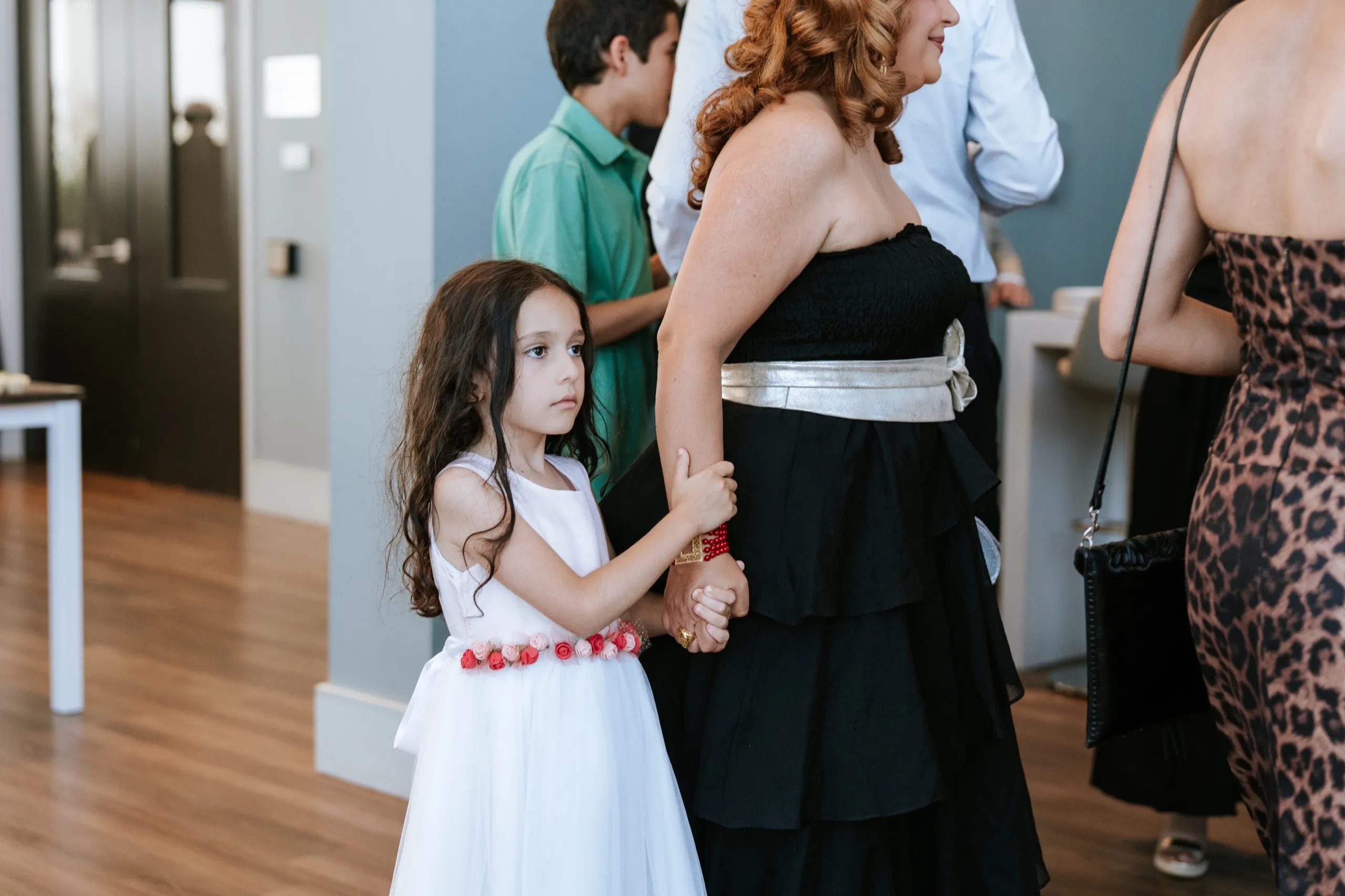 A little girl in a white tulle dress holds on to a family member's hand amid the guests, by AD Photography