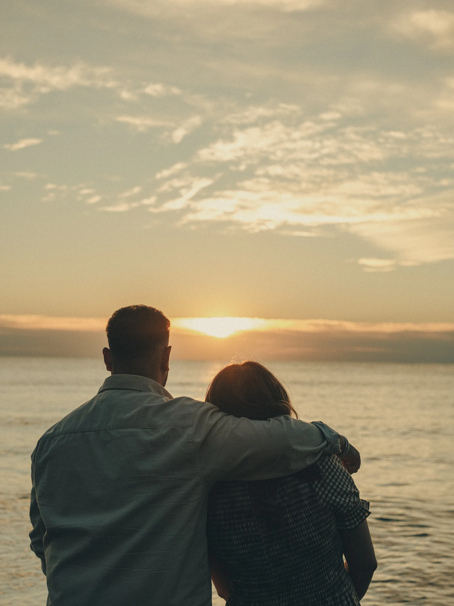 Maninder's arm around Parneet as the sun lifts clear of the low cloud over open water, by AD Photography