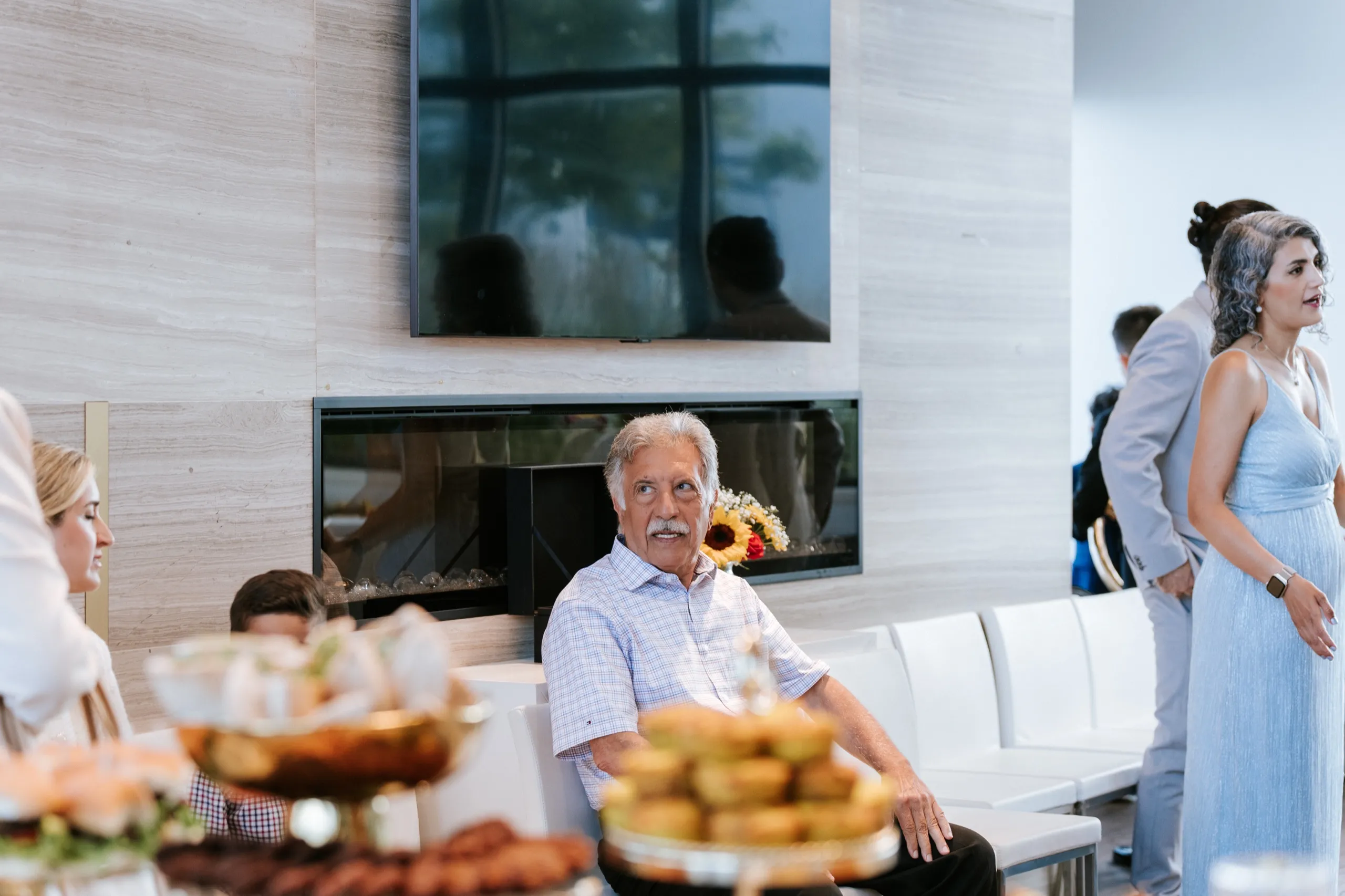 An elder guest looks back from the fireplace lounge, framed past the pastries on the food table, by AD Photography